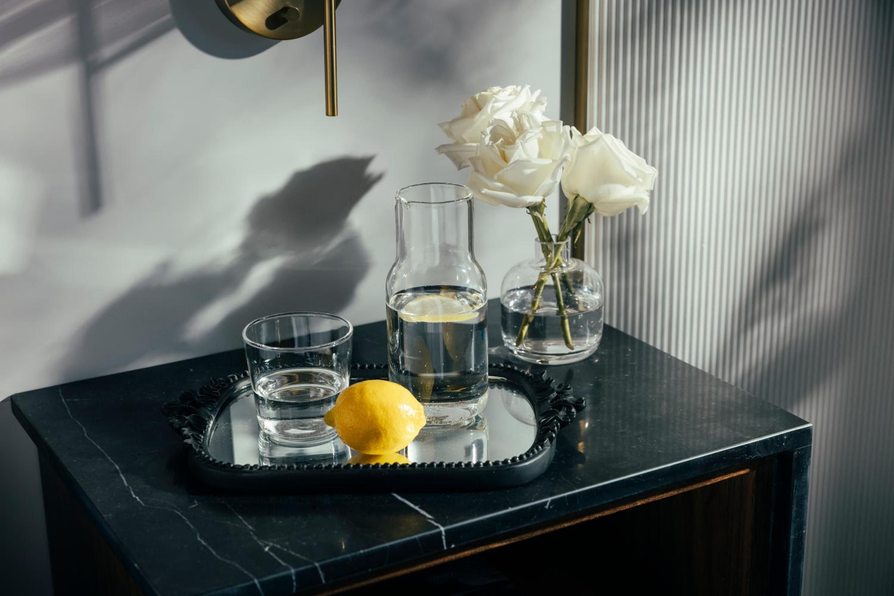 Sunlight streams across marble surfaces, illuminating thoughtful bedside details—fresh white roses, pristine glassware, and a single lemon that speaks to the property's commitment to refined simplicity.