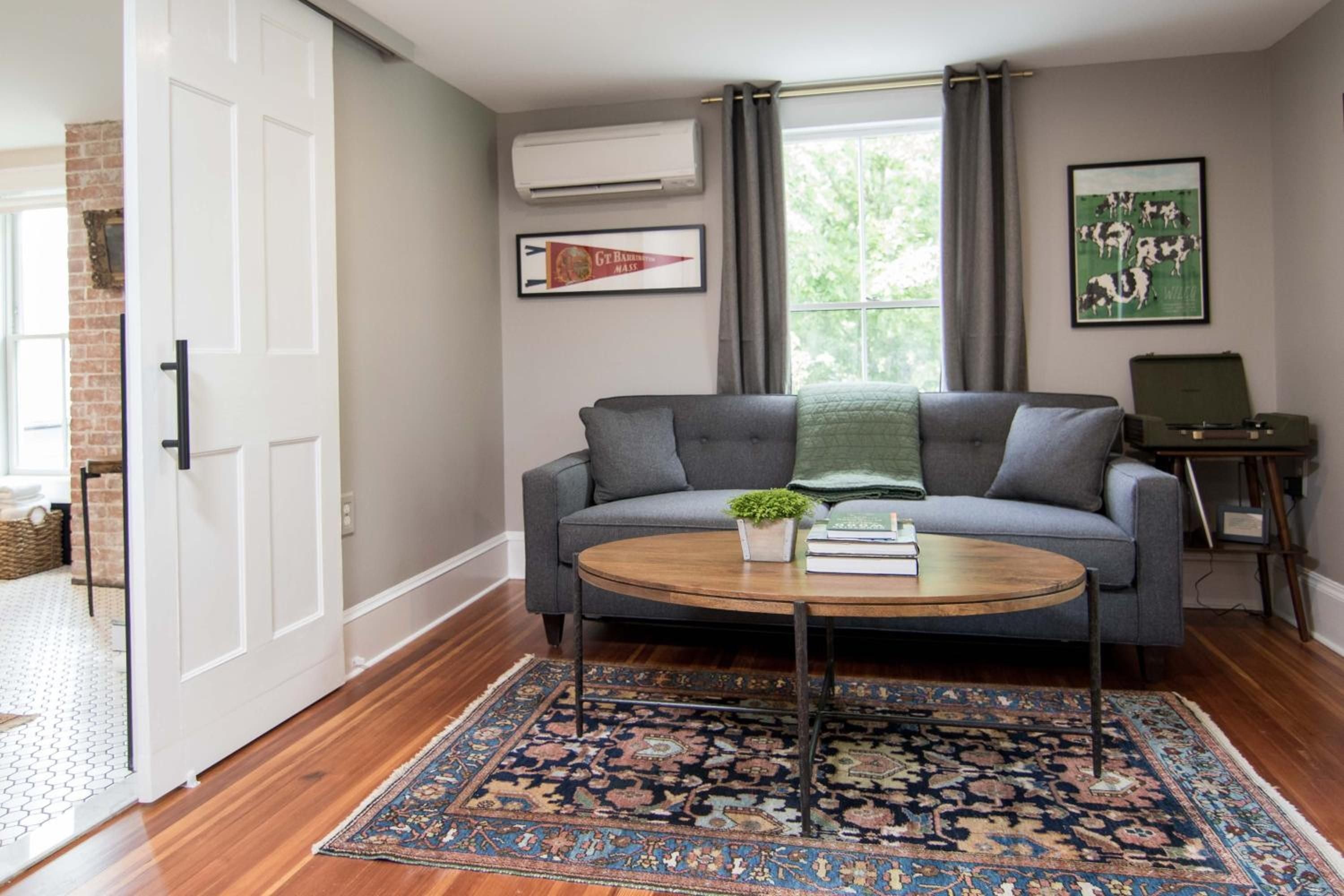 Sunlight filters through gray curtains onto hardwood floors where a Persian rug anchors this inviting living space, complete with vintage pennant and sliding barn door charm.