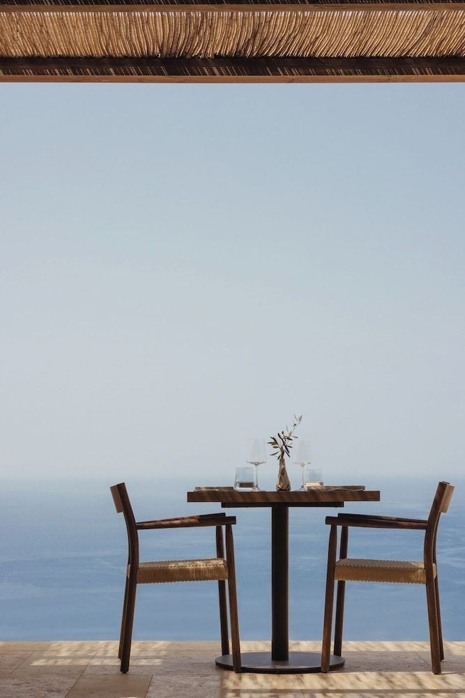 Natural rope fringe creates intimate shade over a perfectly set table for two, where Mediterranean blues stretch endlessly beyond weathered stone floors.