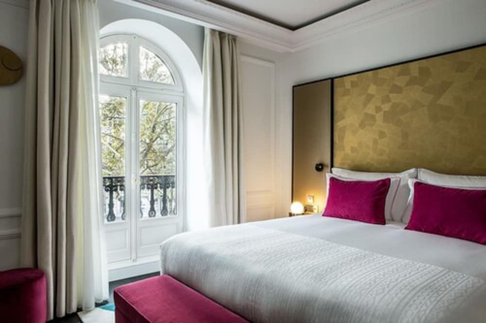 Golden hexagonal headboards and jewel-toned velvet accents create intimate luxury, while arched French doors flood the space with Parisian light through delicate iron balconettes.