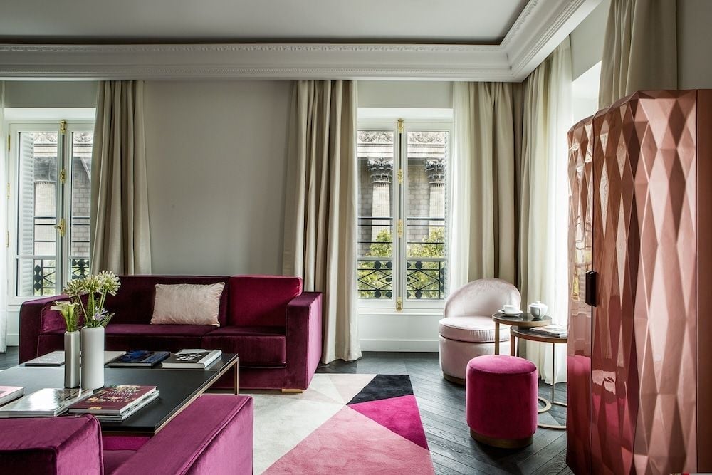 Jewel-toned velvets and geometric copper cabinetry create an opulent Parisian sanctuary, where magenta ottomans and rich burgundy seating frame views of classic Haussmanian architecture.