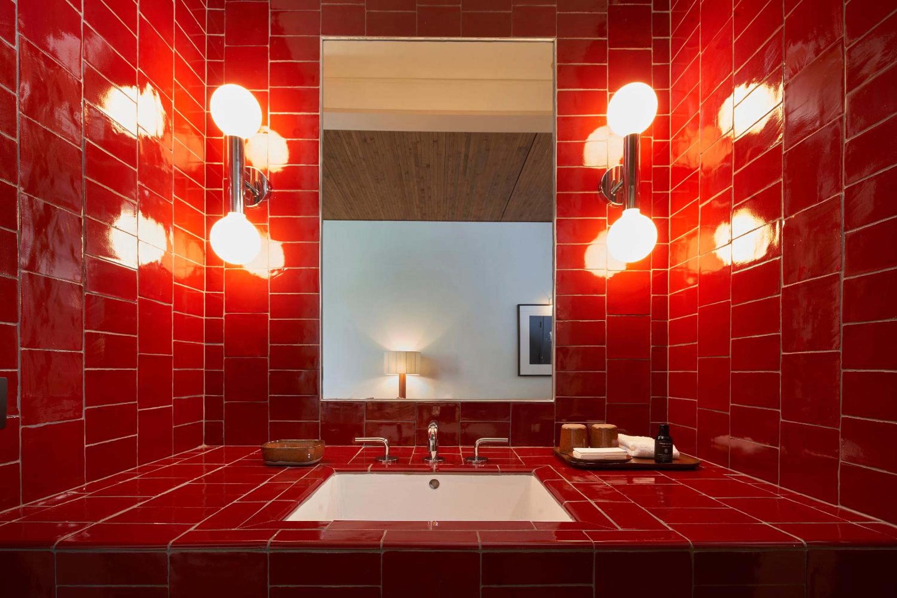 Bold crimson tiles envelop the powder room like a jewel box, where Hollywood-style vanity bulbs cast a theatrical glow over sleek fixtures and warm wood accents.