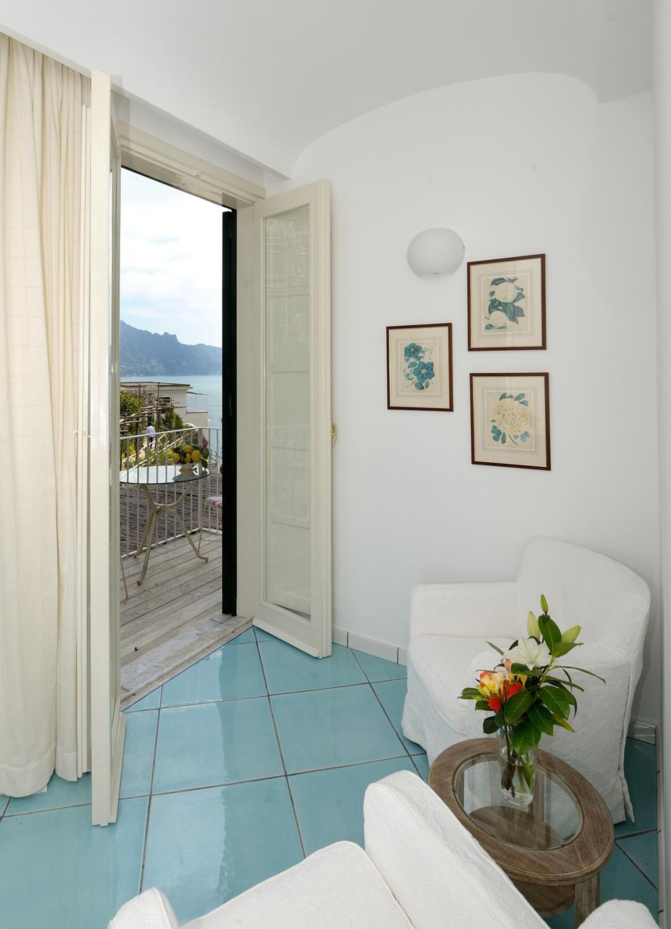 Coastal-inspired blue ceramic floors flow toward French doors revealing the Amalfi's dramatic coastline, while botanical prints add Mediterranean charm to this serene corner.