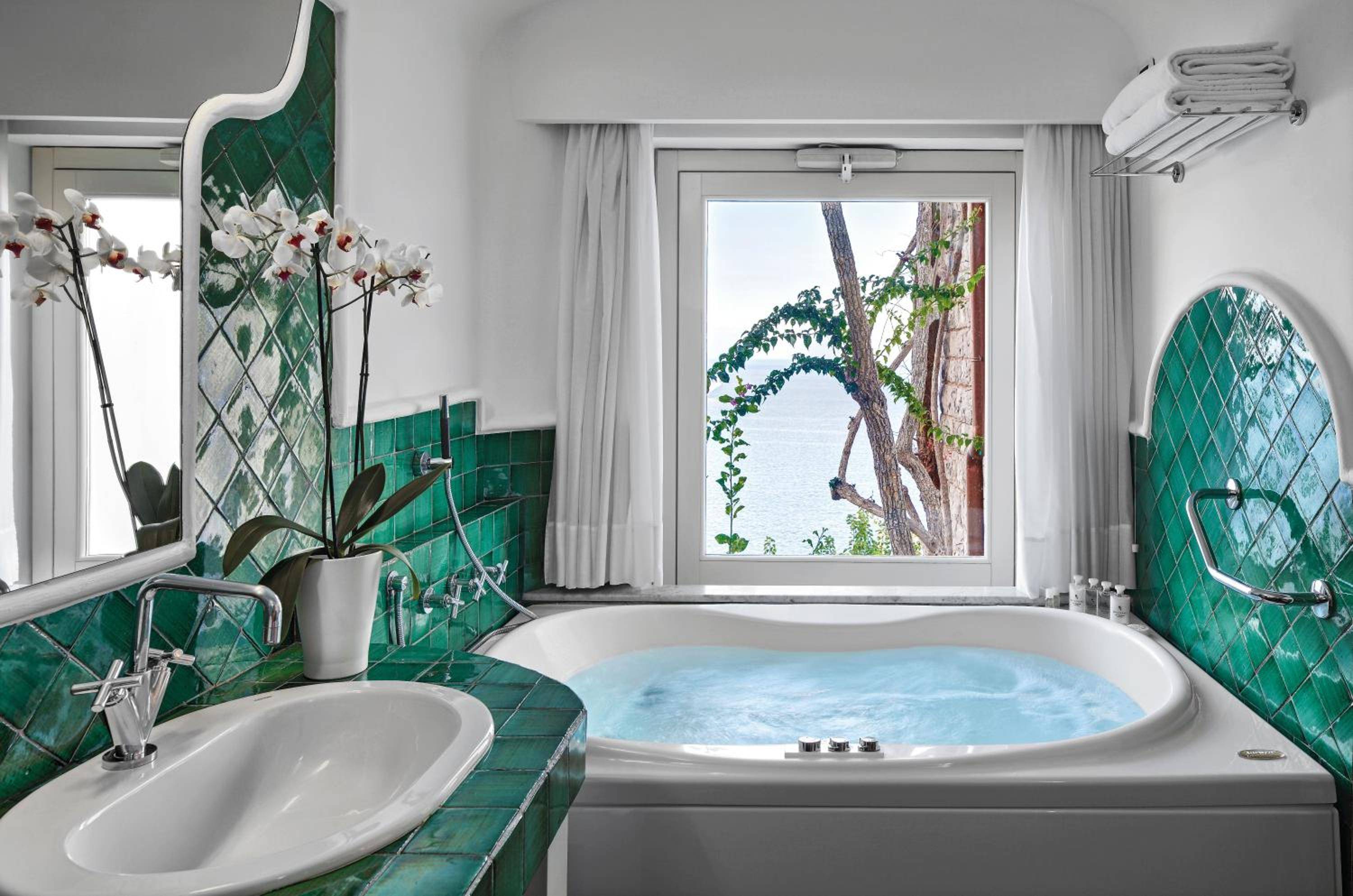 Emerald Majolica tiles frame a marble soaking tub where Mediterranean light filters through windows revealing glimpses of ancient trees and azure coastline beyond.