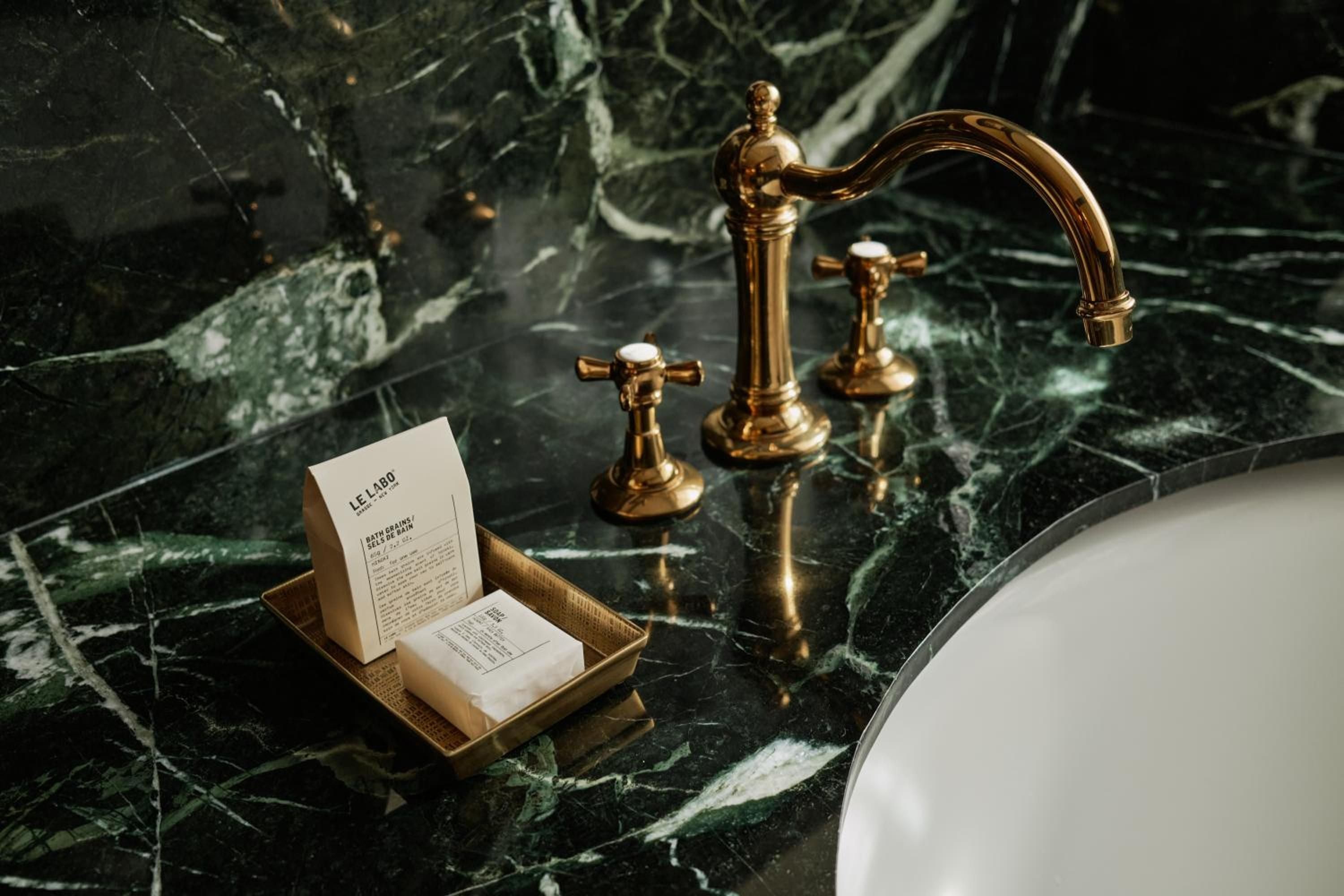 Gleaming brass fixtures catch light against dramatic green marble veining, while artisanal Le Labo amenities transform daily rituals into luxurious moments of indulgence.