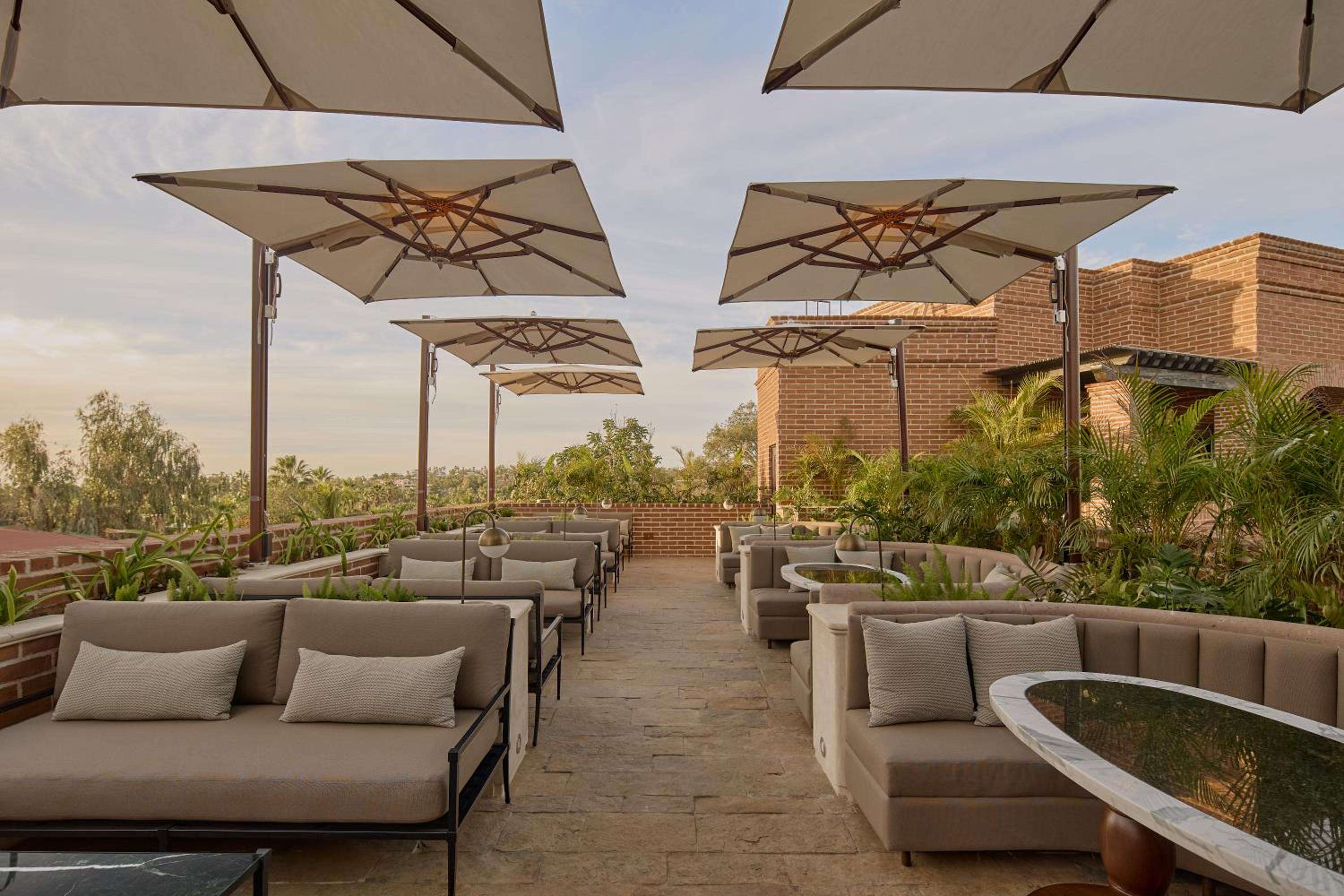 Cantilever umbrellas cast golden shadows across the rooftop terrace, where neutral cushions and curved banquettes create intimate conversation nooks against warm brick architecture.