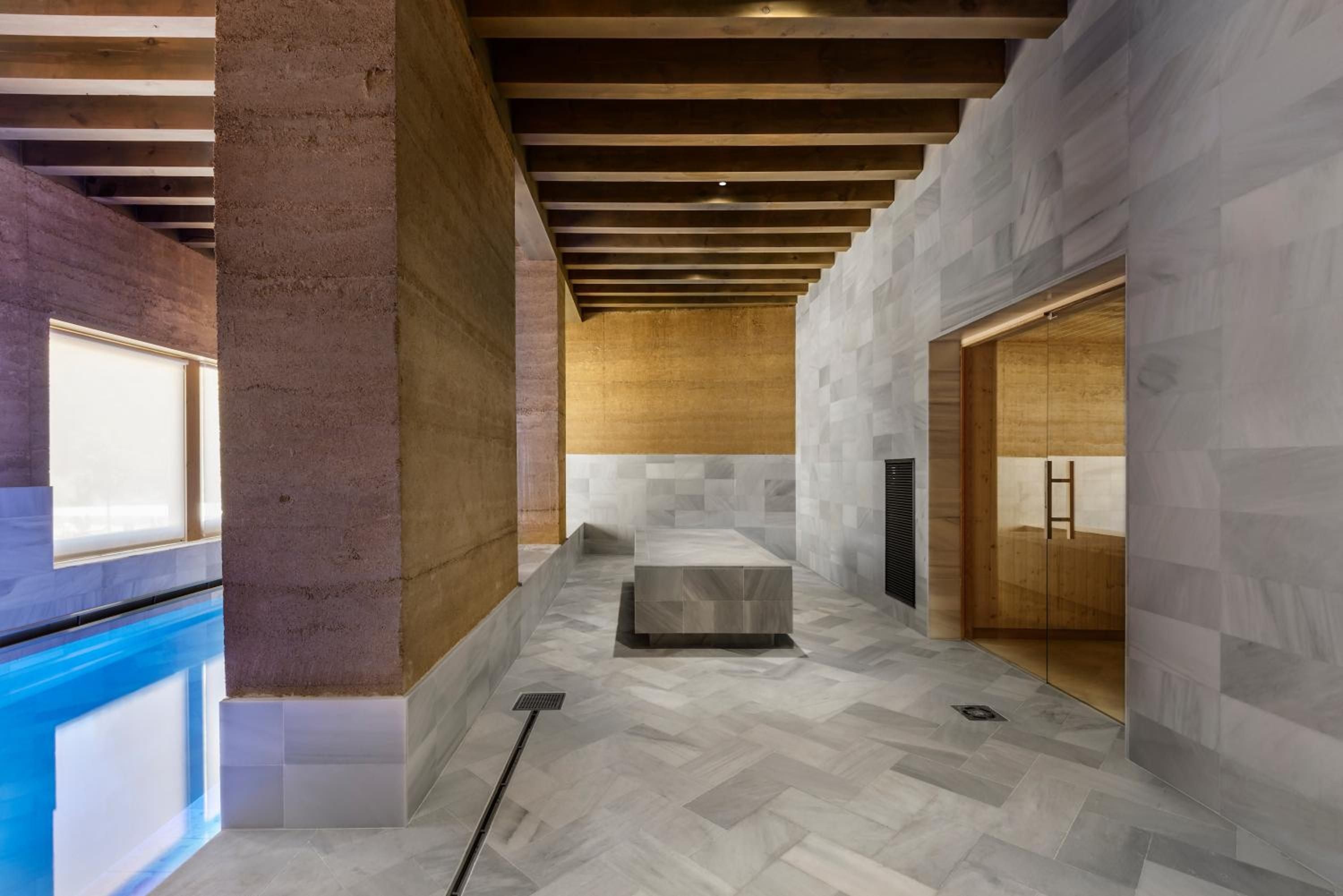 Natural light streams across marble surfaces in the hammam-style spa, where exposed wooden beams frame a serene retreat connecting to the azure indoor pool.