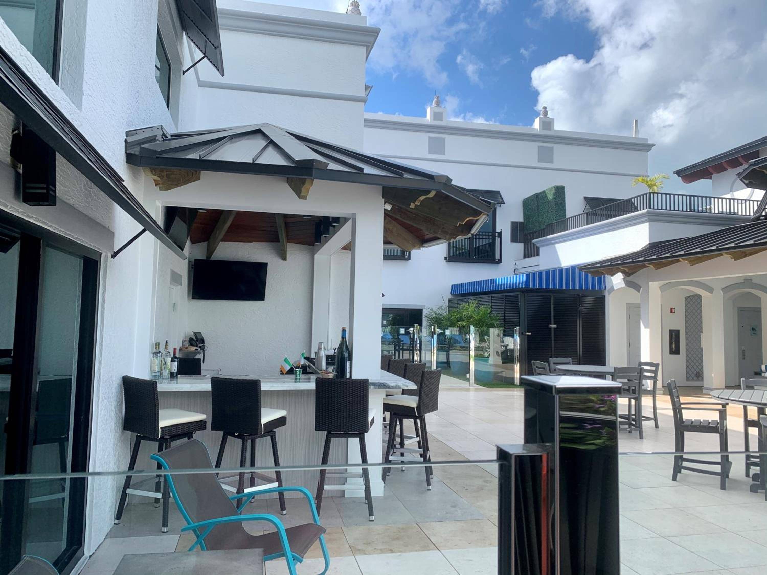 Dark wicker barstools line the open-air rooftop bar where afternoon light filters through sleek metal canopies above pristine white countertops.