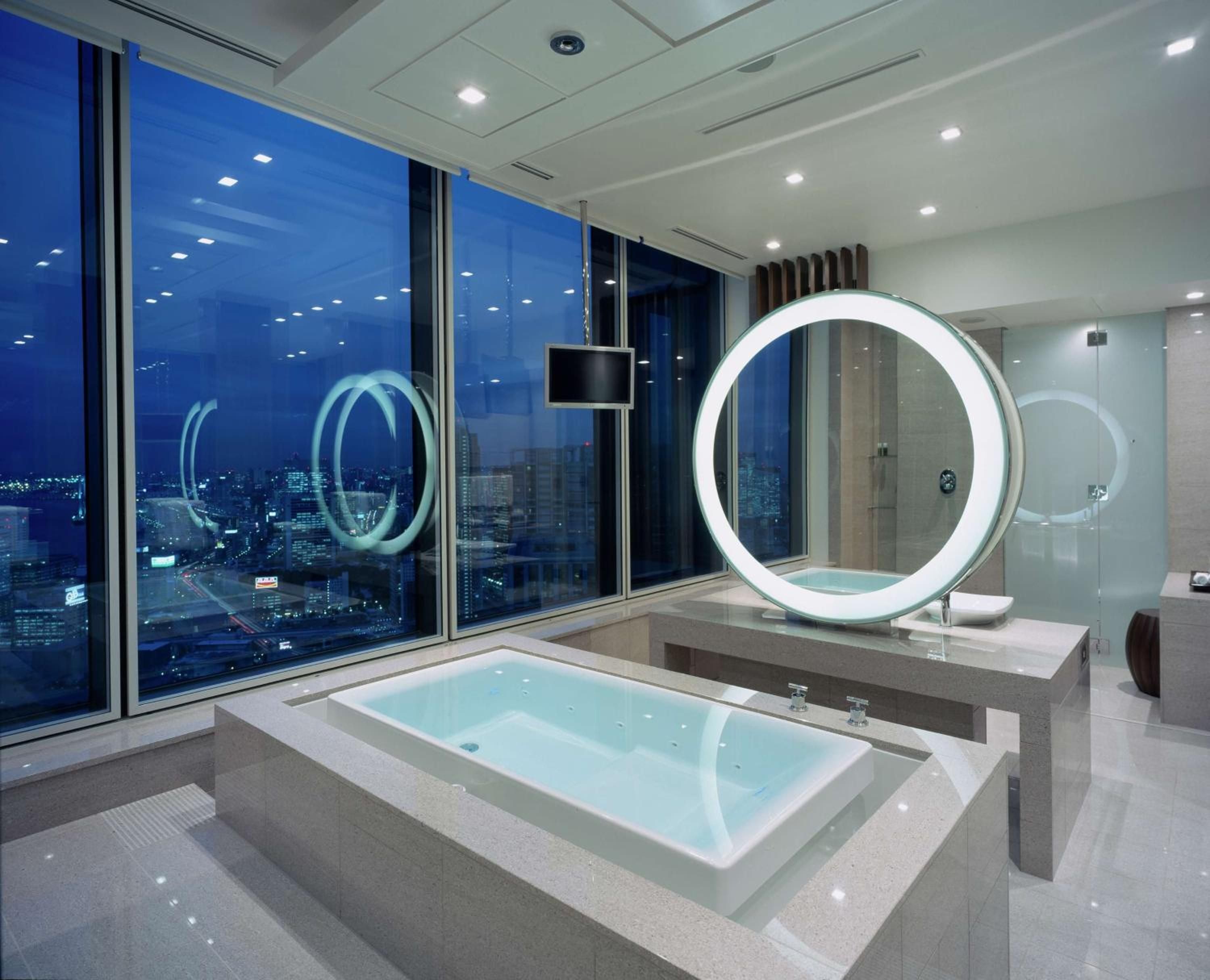 Floor-to-ceiling windows frame Tokyo's glittering skyline while an illuminated circular mirror hovers over the soaking tub like a modern moon in this sky-high sanctuary.
