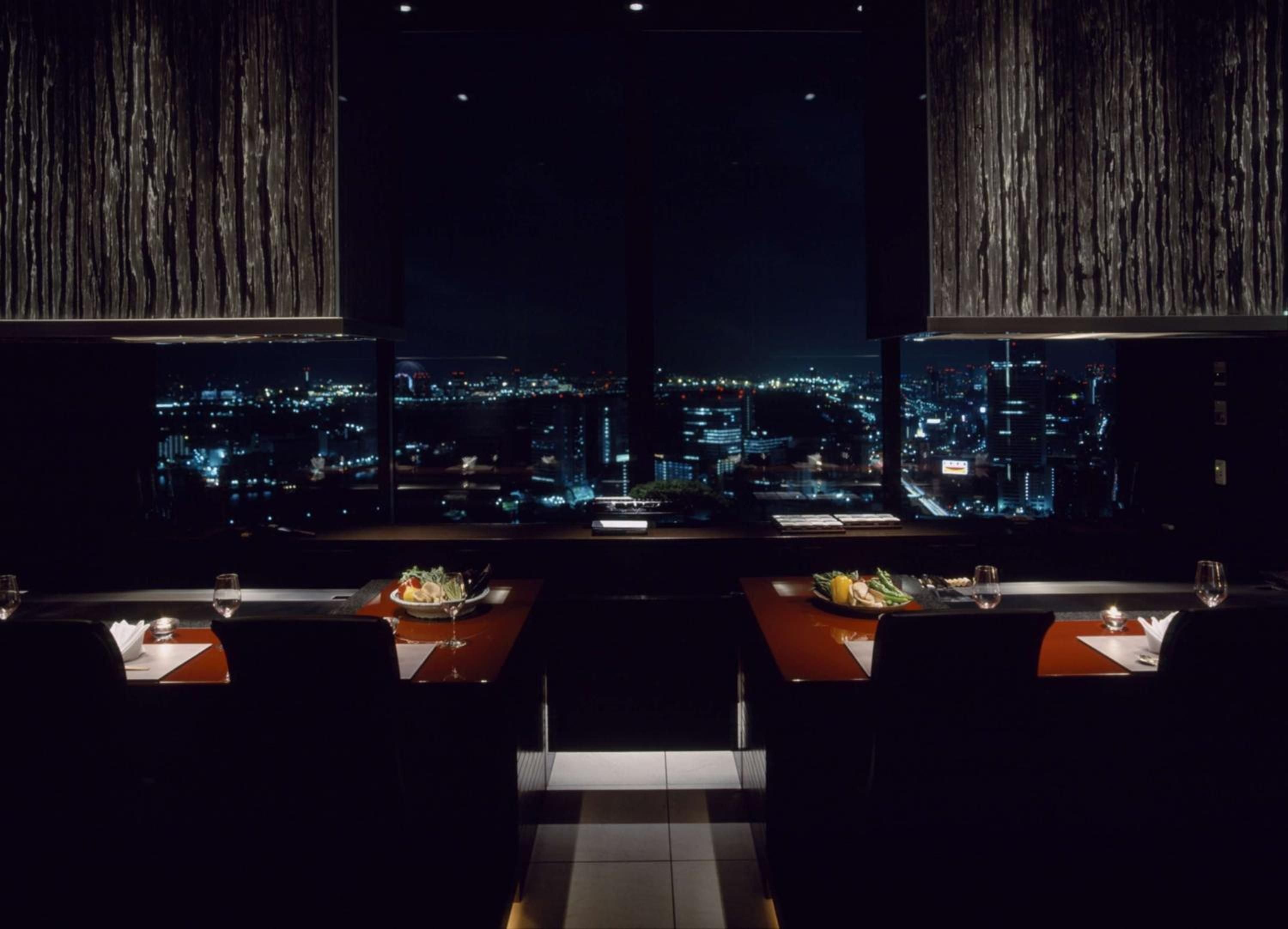 Floor-to-ceiling windows frame Tokyo's glittering skyline while intimate dining tables showcase artful kaiseki presentations against warm wood textures and dramatic low lighting.
