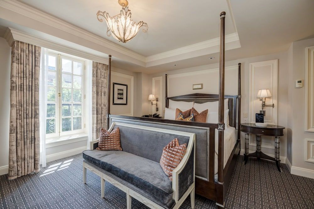 Soft afternoon light filters through tall windows into this sophisticated guest room, where a four-poster bed and velvet settee create an intimate retreat with timeless elegance.