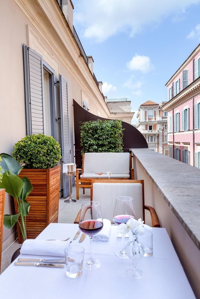 Al fresco dining unfolds on a sun-drenched terrace where crisp linens flutter against Rome's pastel-hued rooftops and azure Mediterranean sky.