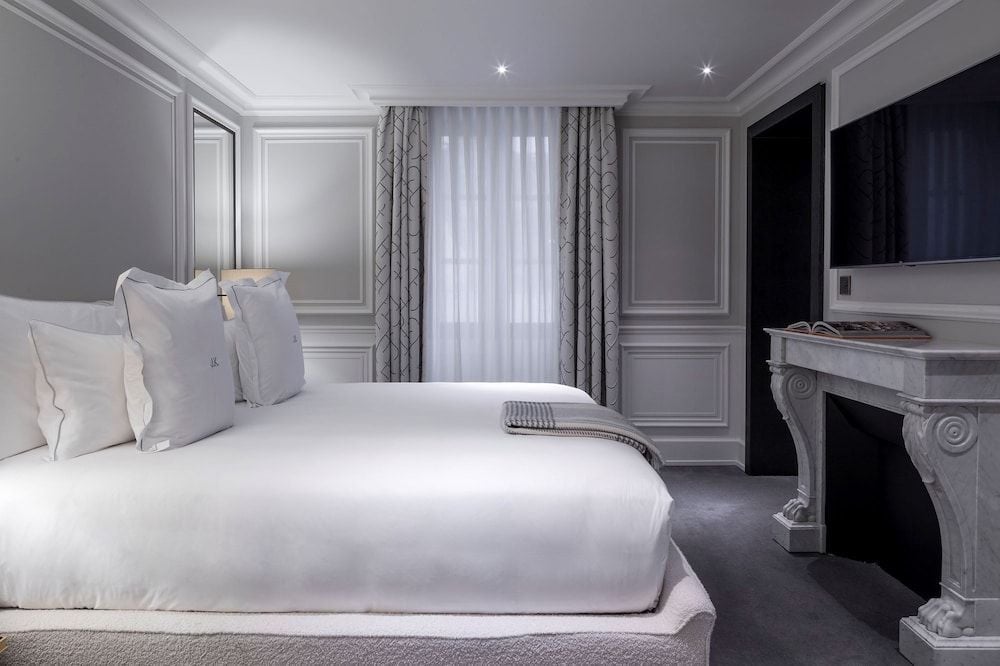 Parisian elegance meets understated luxury in this serene guest room, where dove-gray moldings frame crisp white linens and geometric curtains filter afternoon light through tall windows.