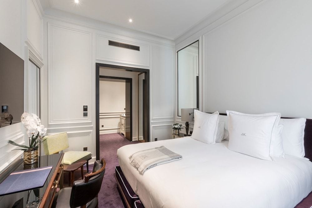 Crisp white linens meet rich burgundy carpets in this elegantly appointed suite, where classical Parisian moldings frame modern comforts and soft recessed lighting creates intimate ambiance.