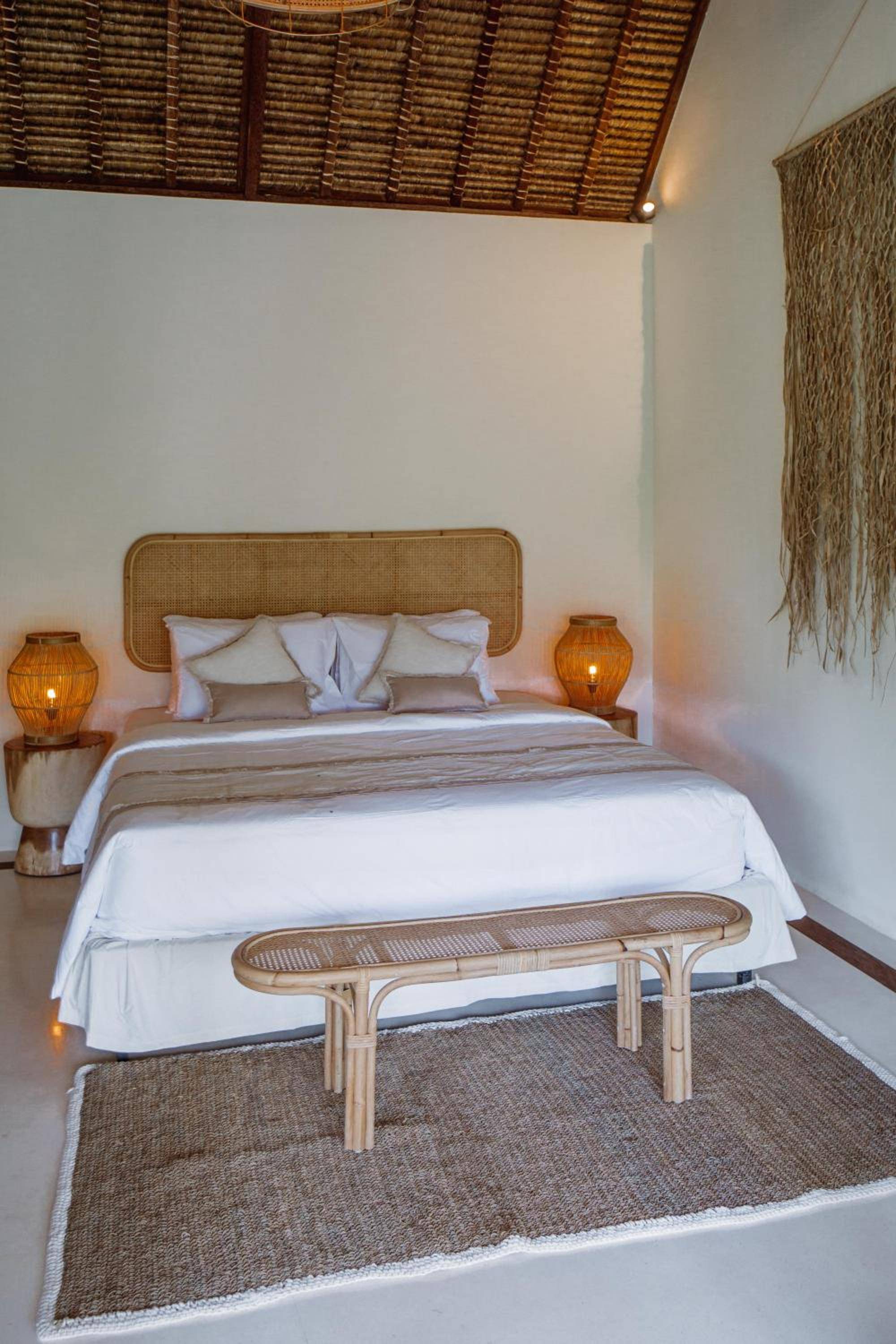 Natural rattan headboards and woven ceiling details create an organic sanctuary, while amber pendant lamps cast honeyed light across crisp white linens and jute textures.