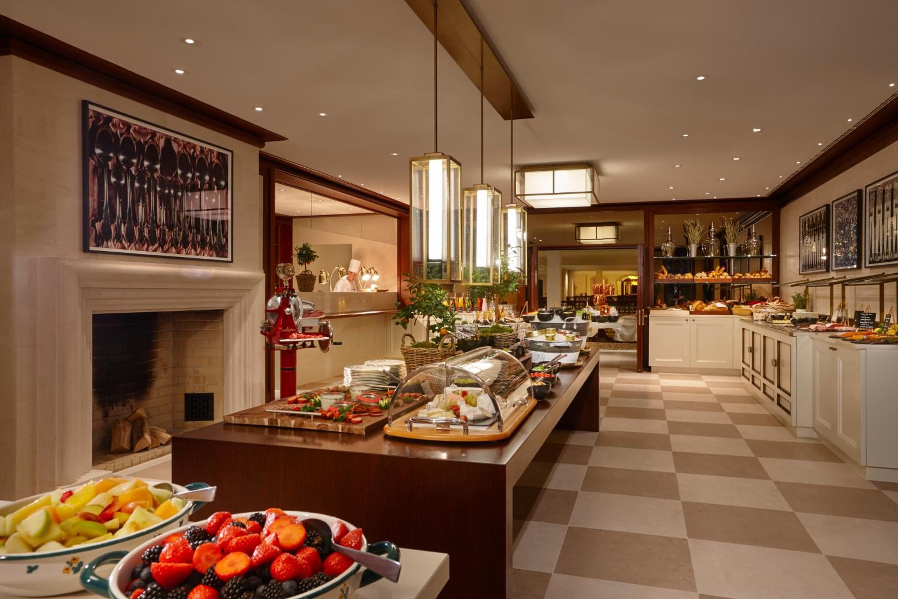 Marble floors gleam beneath pendant lights as the breakfast buffet unfolds across polished counters, while flames flicker in the grand fireplace beyond.