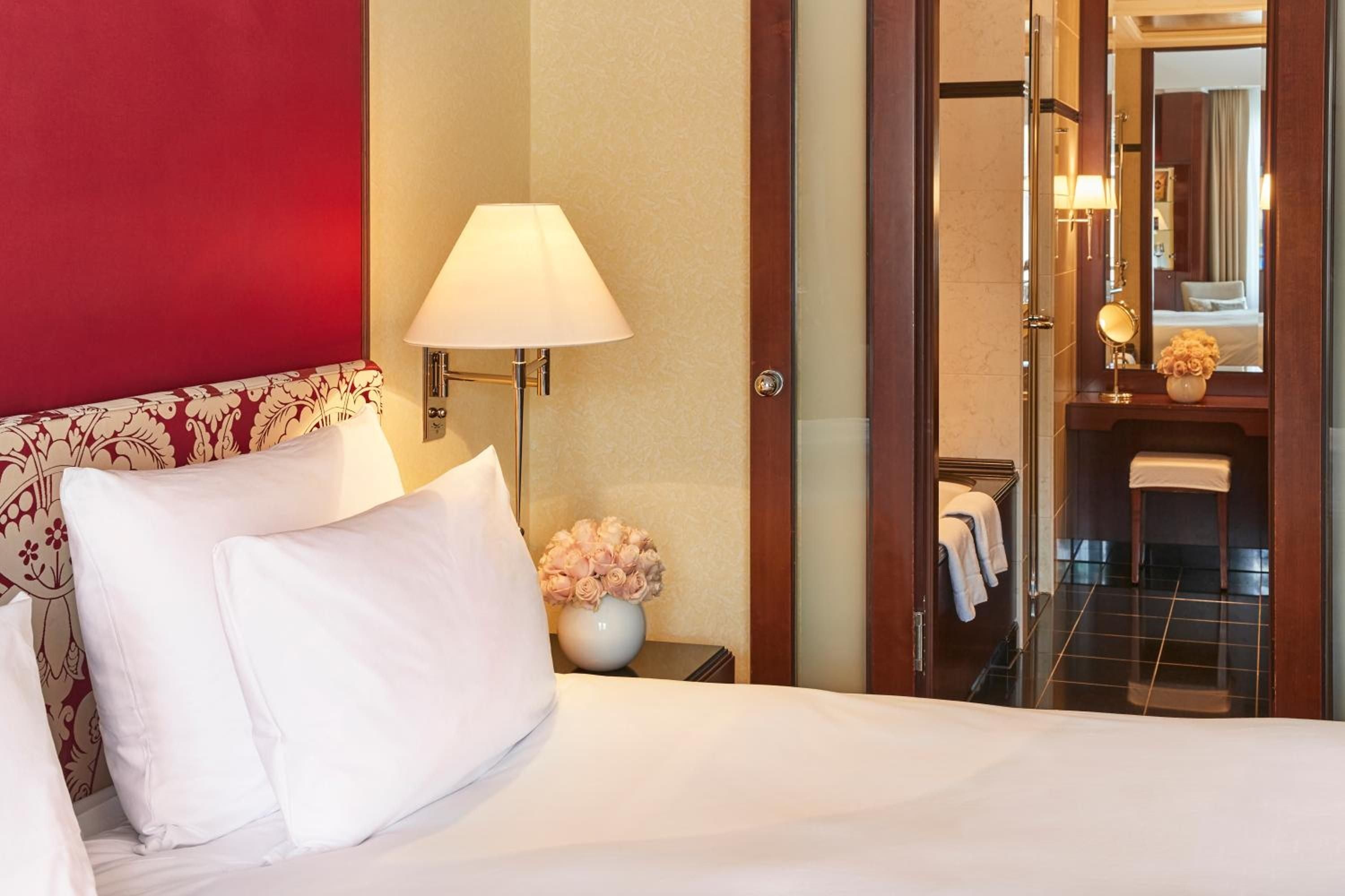 Crisp white linens contrast beautifully with burgundy floral headboard details, while warm lamplight and fresh roses create an intimate, sophisticated ambiance in this elegantly appointed guest room.