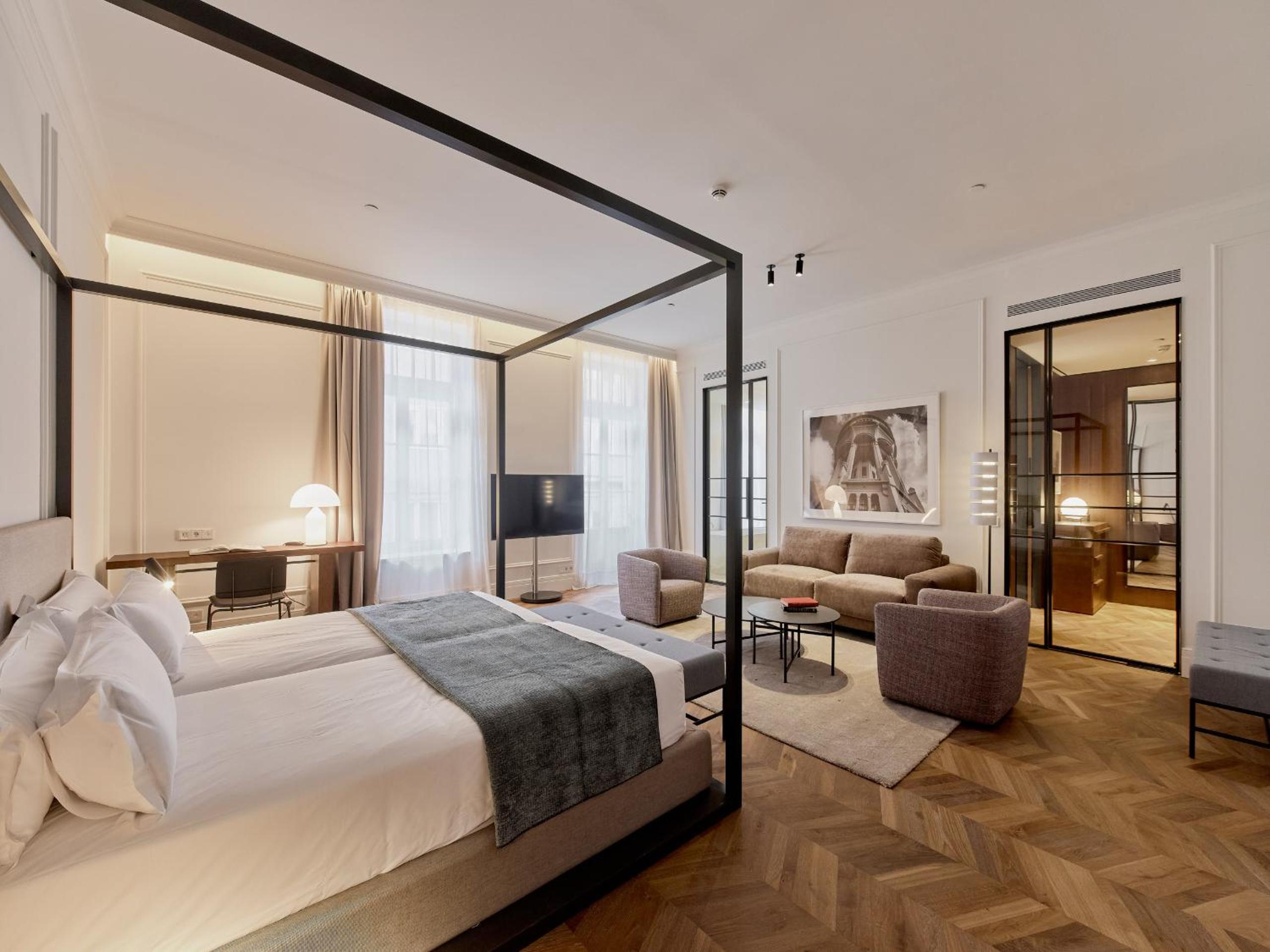 Herringbone parquet floors gleam beneath a sleek canopy bed, while neutral fabrics and black steel frames create sophisticated harmony in this airy Budapest suite.