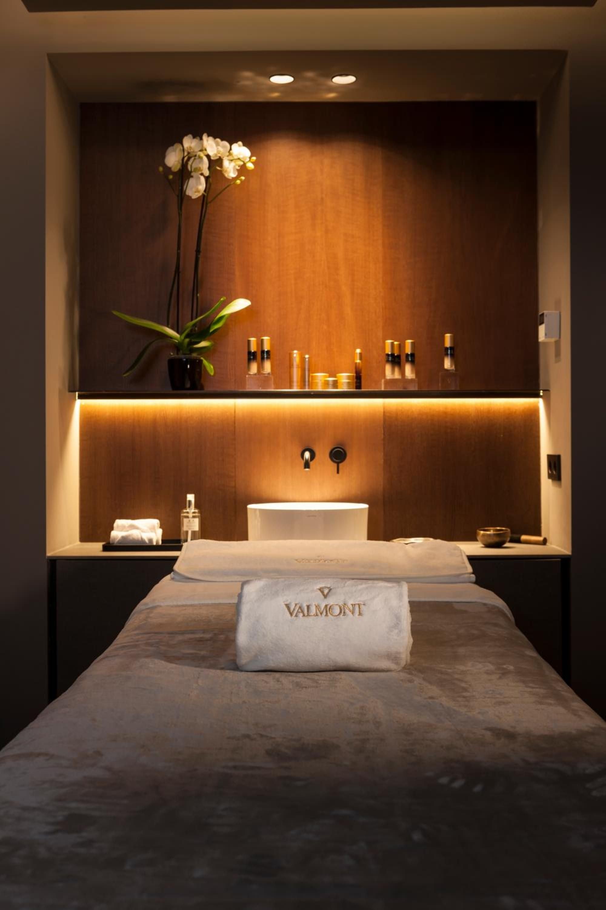 Warm amber lighting bathes the wood-paneled treatment room where Valmont linens await, while orchids and precisely arranged skincare products create an intimate wellness sanctuary.
