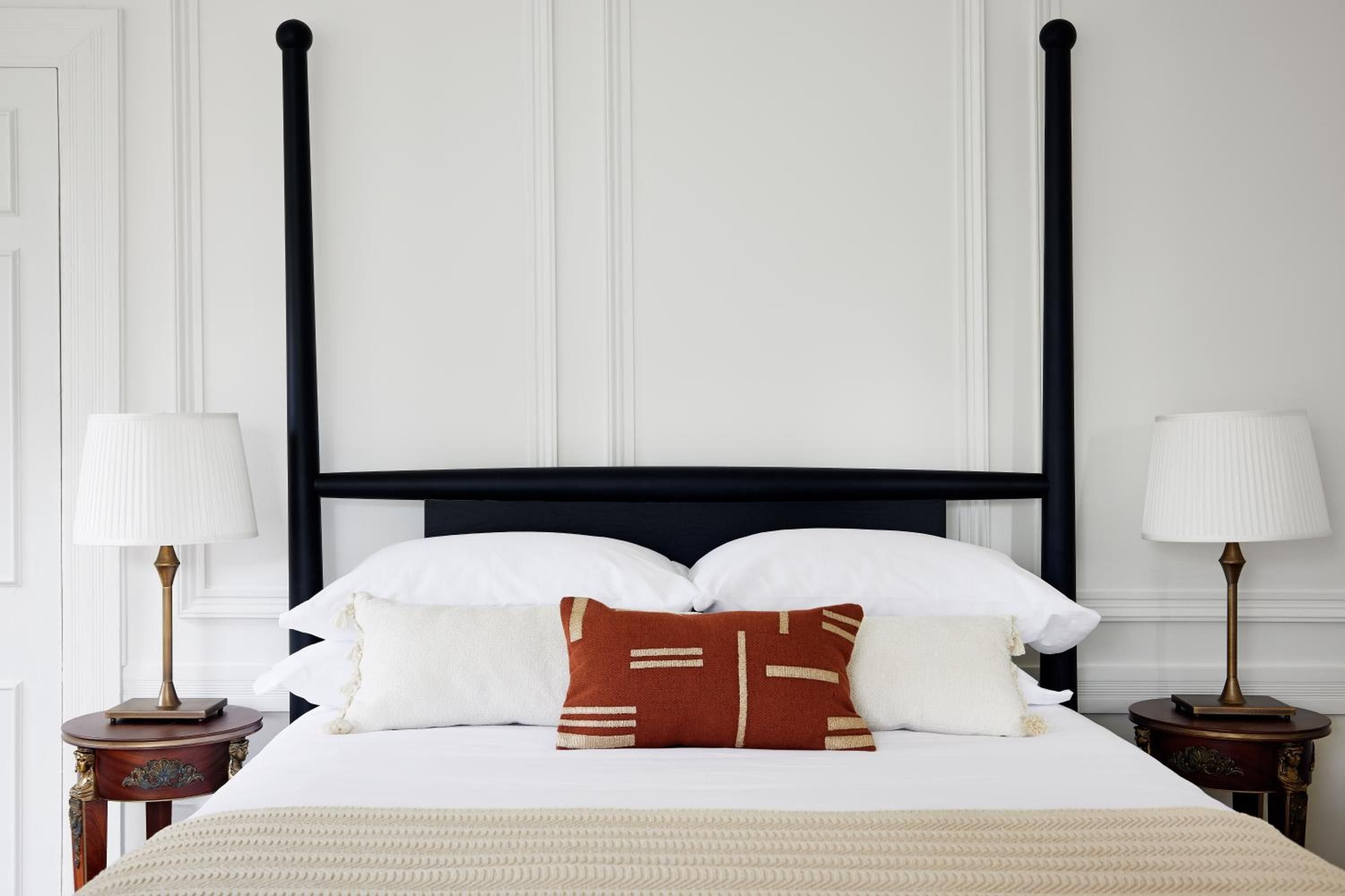 Crisp white linens contrast beautifully with the black four-poster bed frame, while warm brass lamps and a rust-colored accent pillow create intimate sophistication.