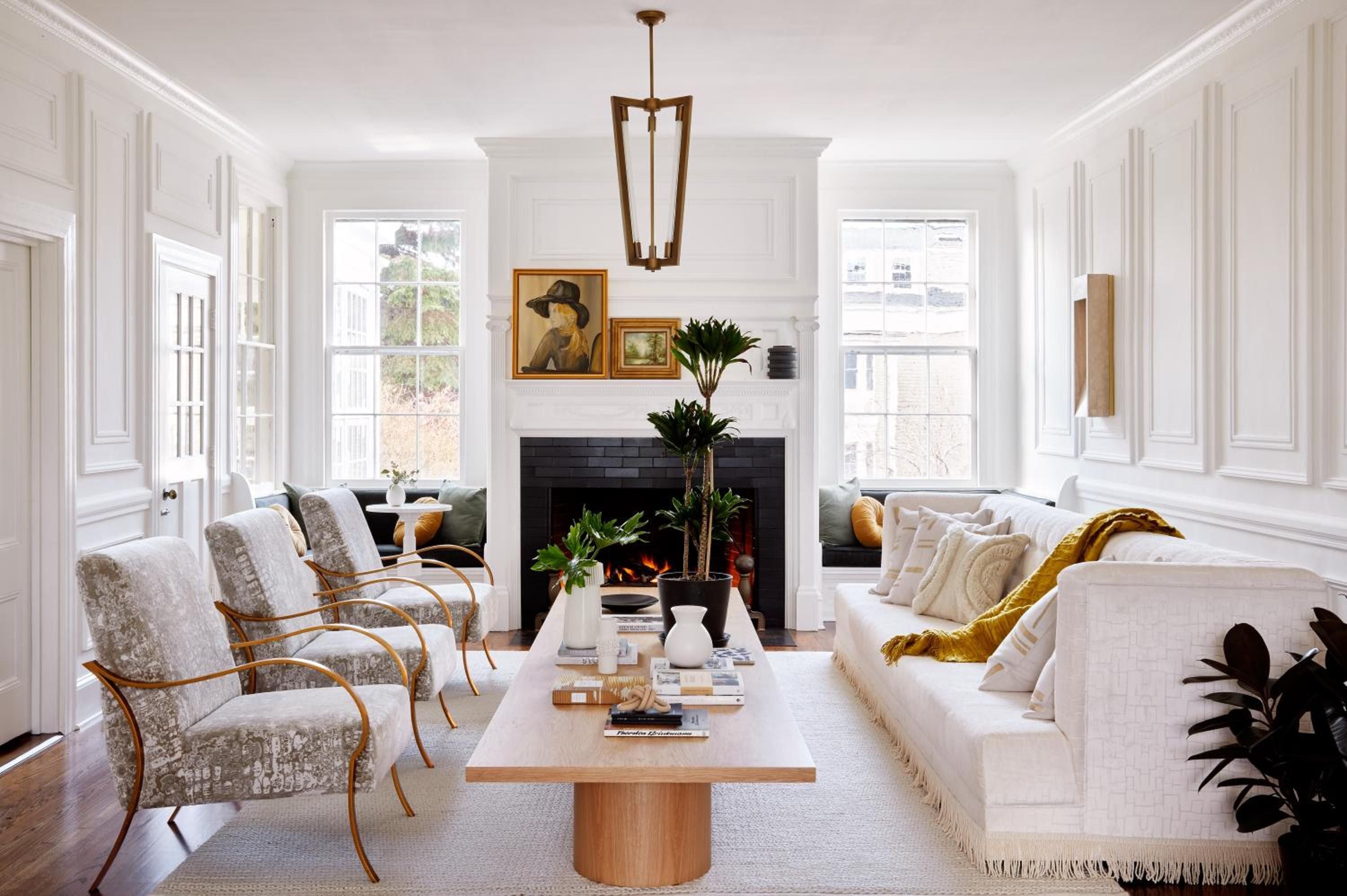 Velvet armchairs with brass frames face a crackling fireplace, while afternoon light streams through tall windows onto marble surfaces and carefully curated botanical arrangements.