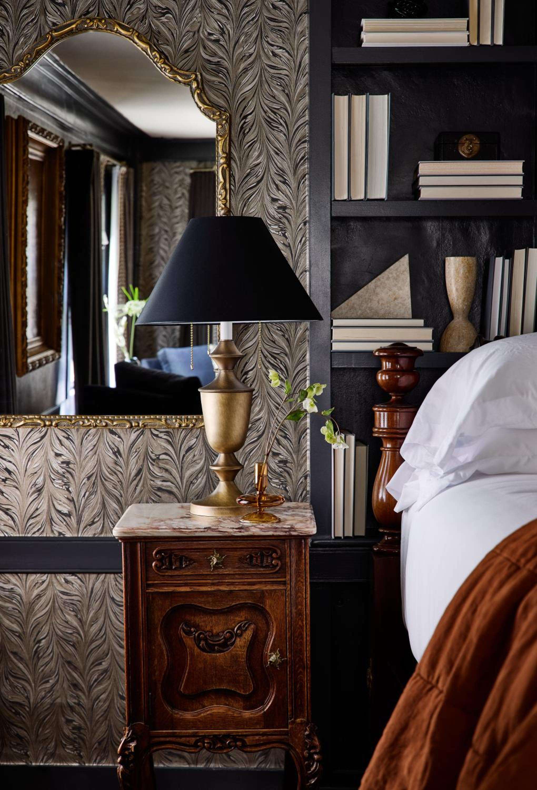 Ornate wallpaper meets curated bookshelves in this thoughtfully appointed guest room, where brass fixtures and carved wood nightstands create an intimate literary sanctuary.