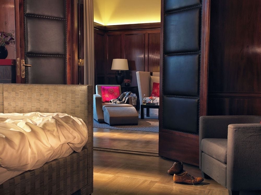 Rich mahogany doors frame glimpses of the opulent sitting area, where warm lamplight spills across plush furnishings and leather-bound luxury awaits beyond woven headboard textures.