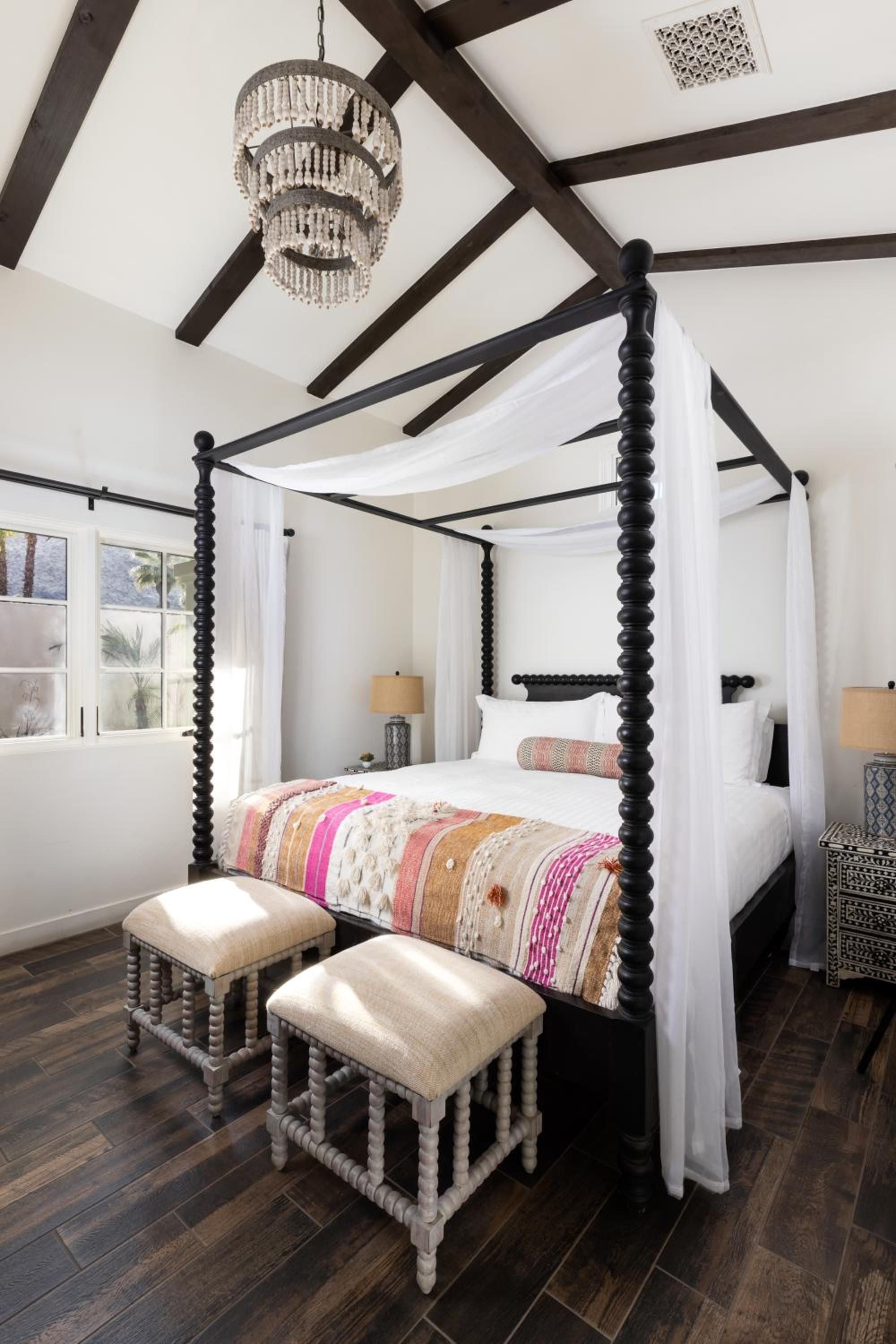 Exposed wooden beams frame a dramatic four-poster bed adorned with vibrant Mexican textiles, while crystal chandeliers cast warm light across dark hardwood floors.