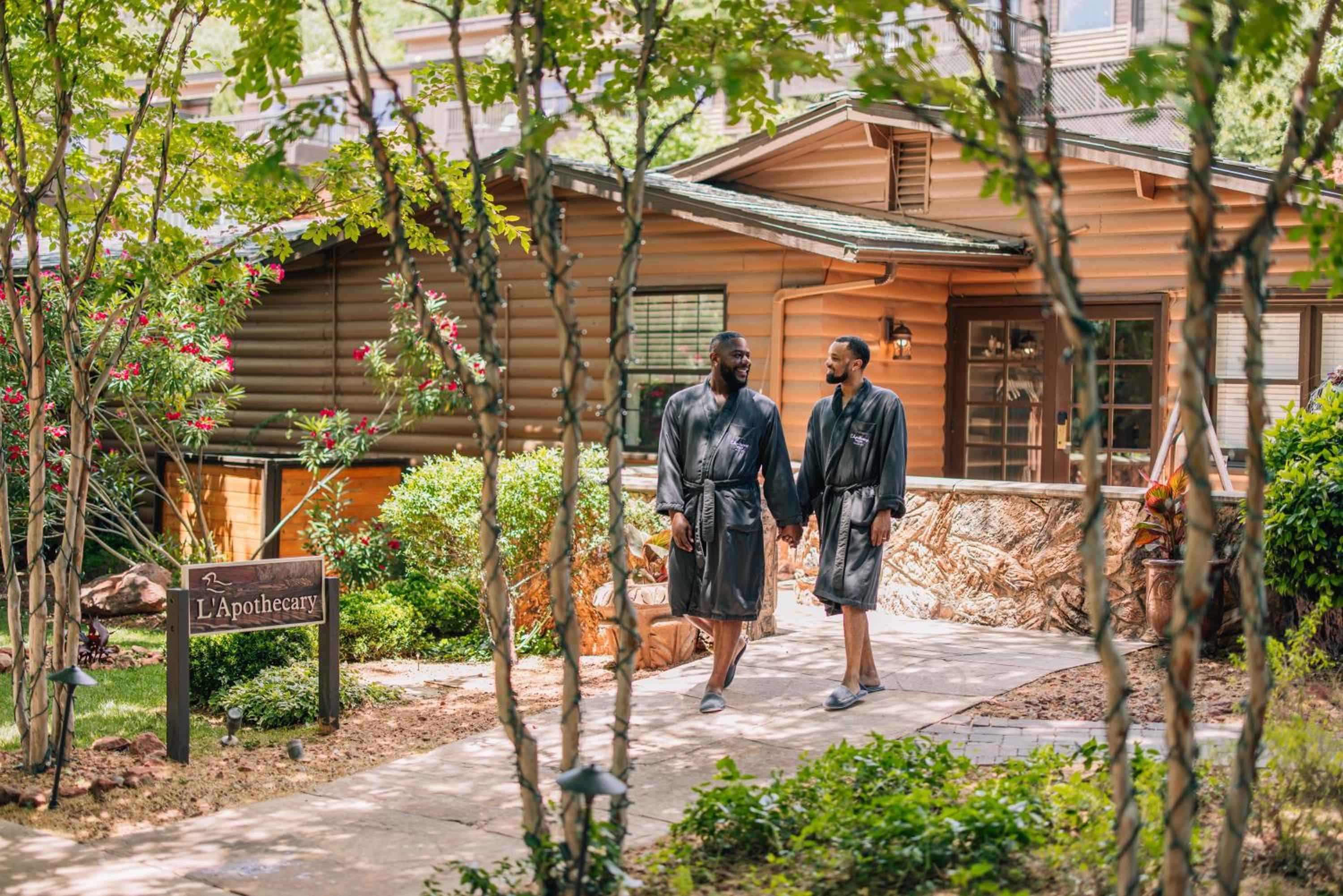 Two guests in plush robes stroll along stone pathways toward L'Apothecary spa, where rustic log cabins nestle among flowering shrubs and dappled forest light.