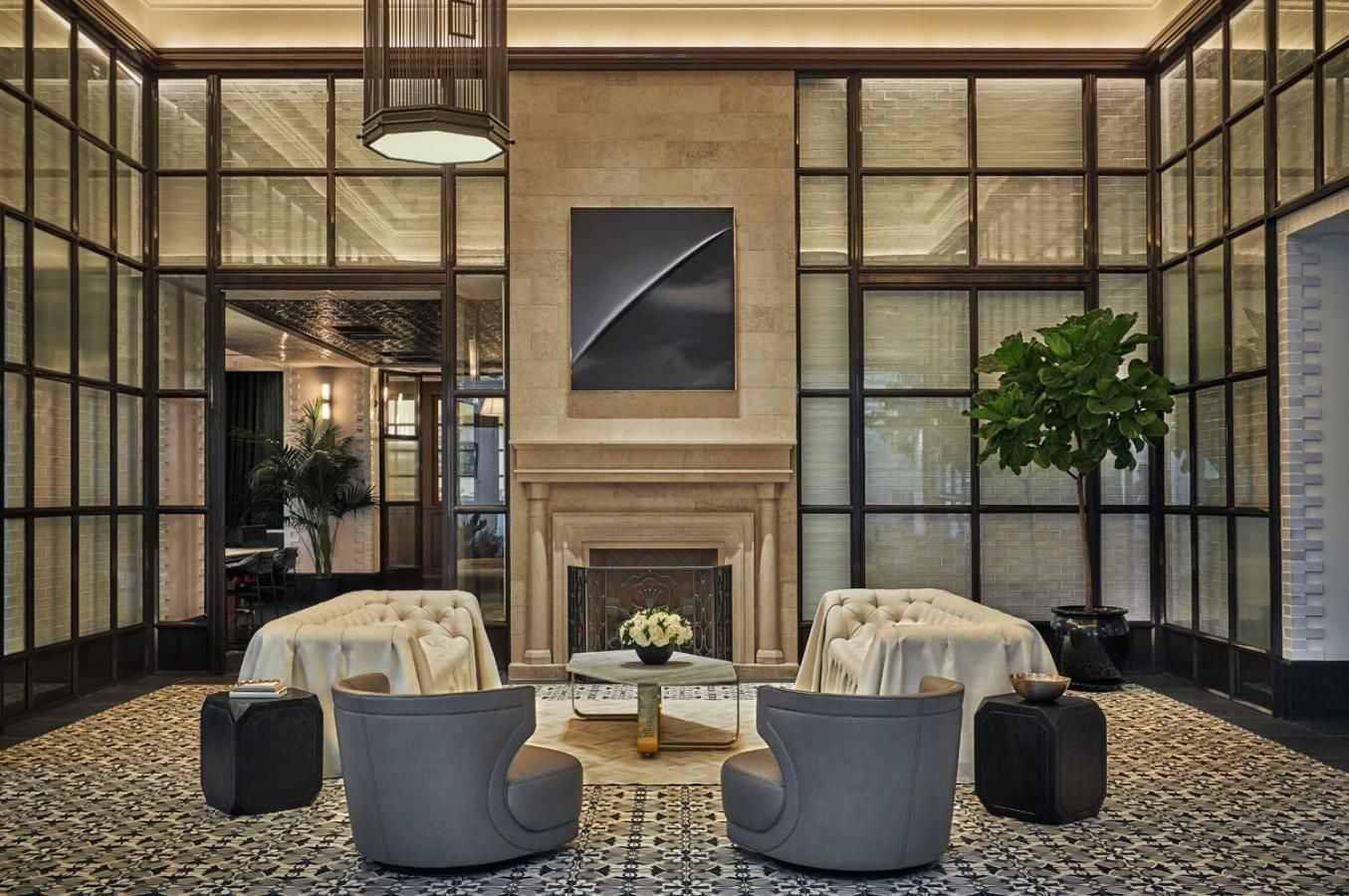 Floor-to-ceiling steel-framed windows flood the sophisticated lobby with natural light, where plush swivel chairs invite intimate conversations beside the limestone fireplace.