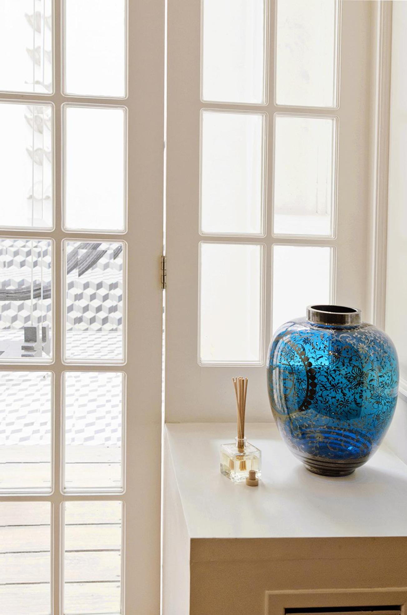 Sunlight filters through French doors onto a marble surface where an ornate blue ceramic vase and crystal reed diffuser create an intimate vignette of refined Mexican elegance.