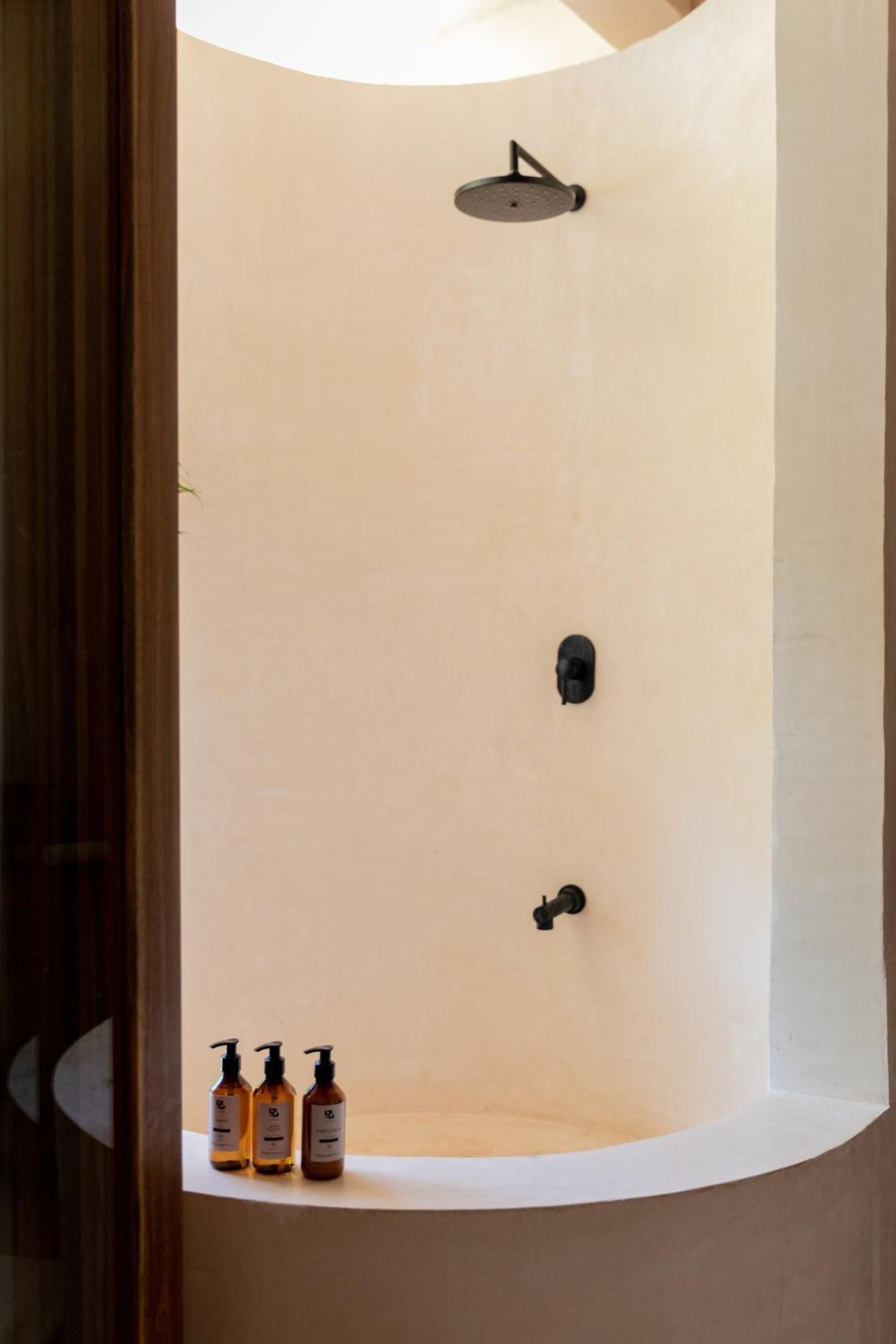 Curved white walls embrace a rainfall shower where amber-bottled amenities rest on the ledge, creating a spa-like sanctuary with modern black fixtures.