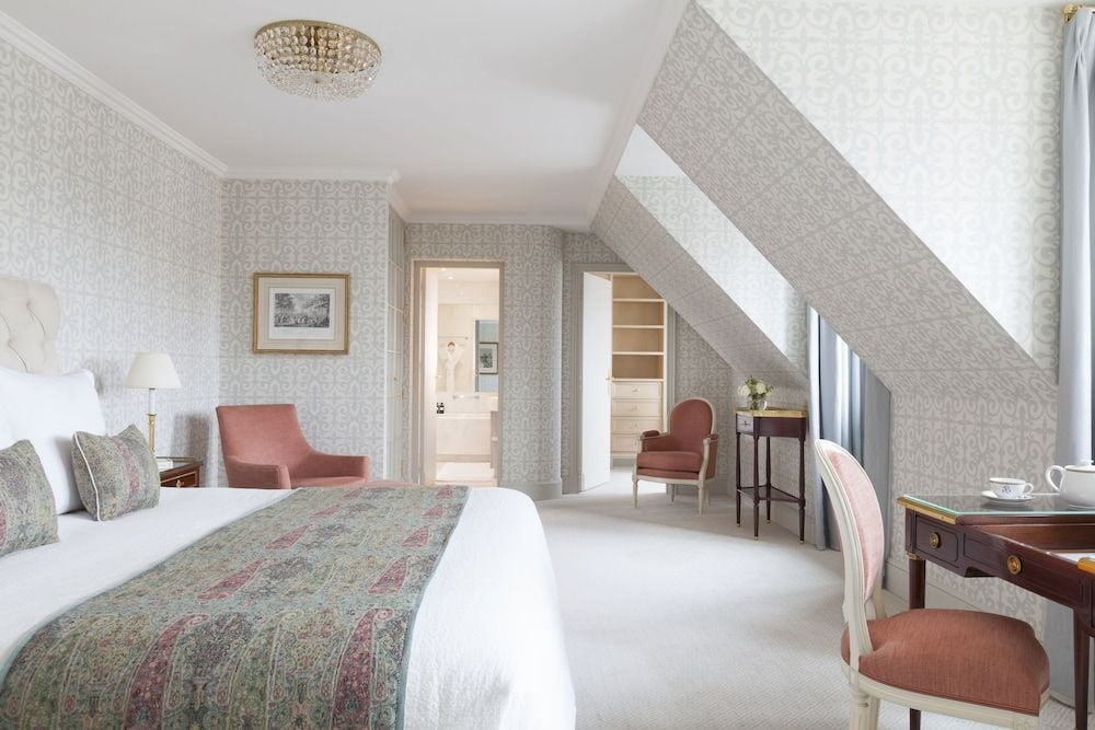 Soft gray damask wallpaper and coral-hued furnishings create an intimate Parisian retreat beneath slanted rooflines, where crystal chandeliers cast gentle light across luxurious linens.