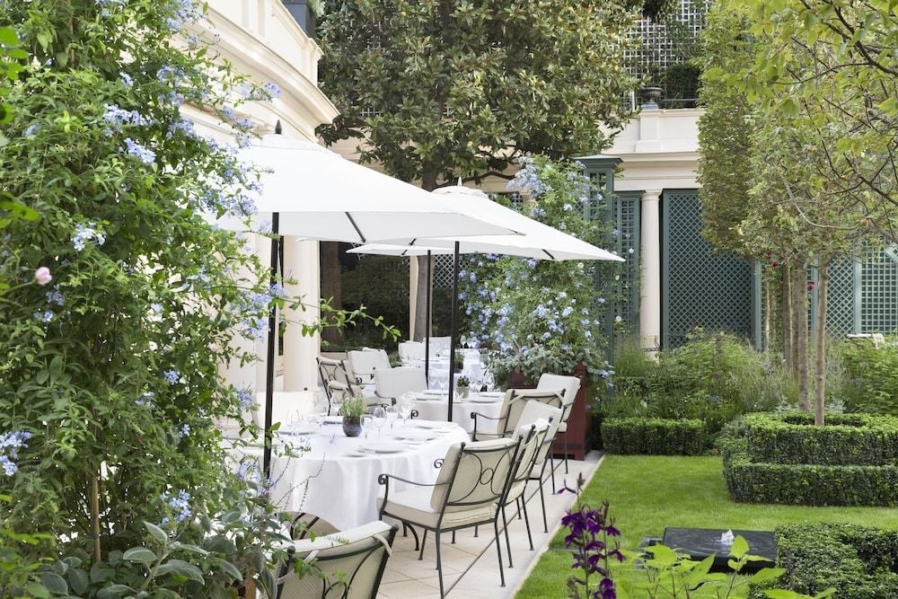 White umbrellas shade intimate tables on the terrace, where blue plumbago blooms frame perfectly manicured hedgerows and classical French architecture creates an enchanting dining sanctuary.