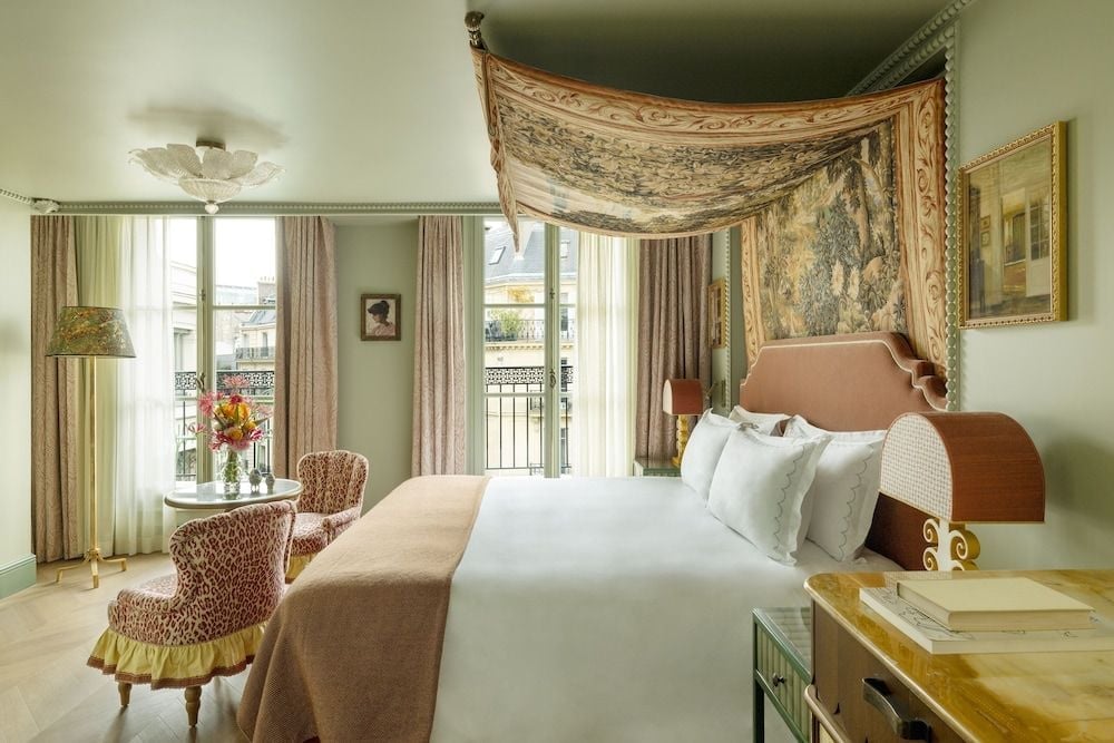 Sunlight streams through tall French windows onto crisp linens and an ornate tapestry canopy, where Parisian elegance meets intimate luxury in this thoughtfully appointed suite.