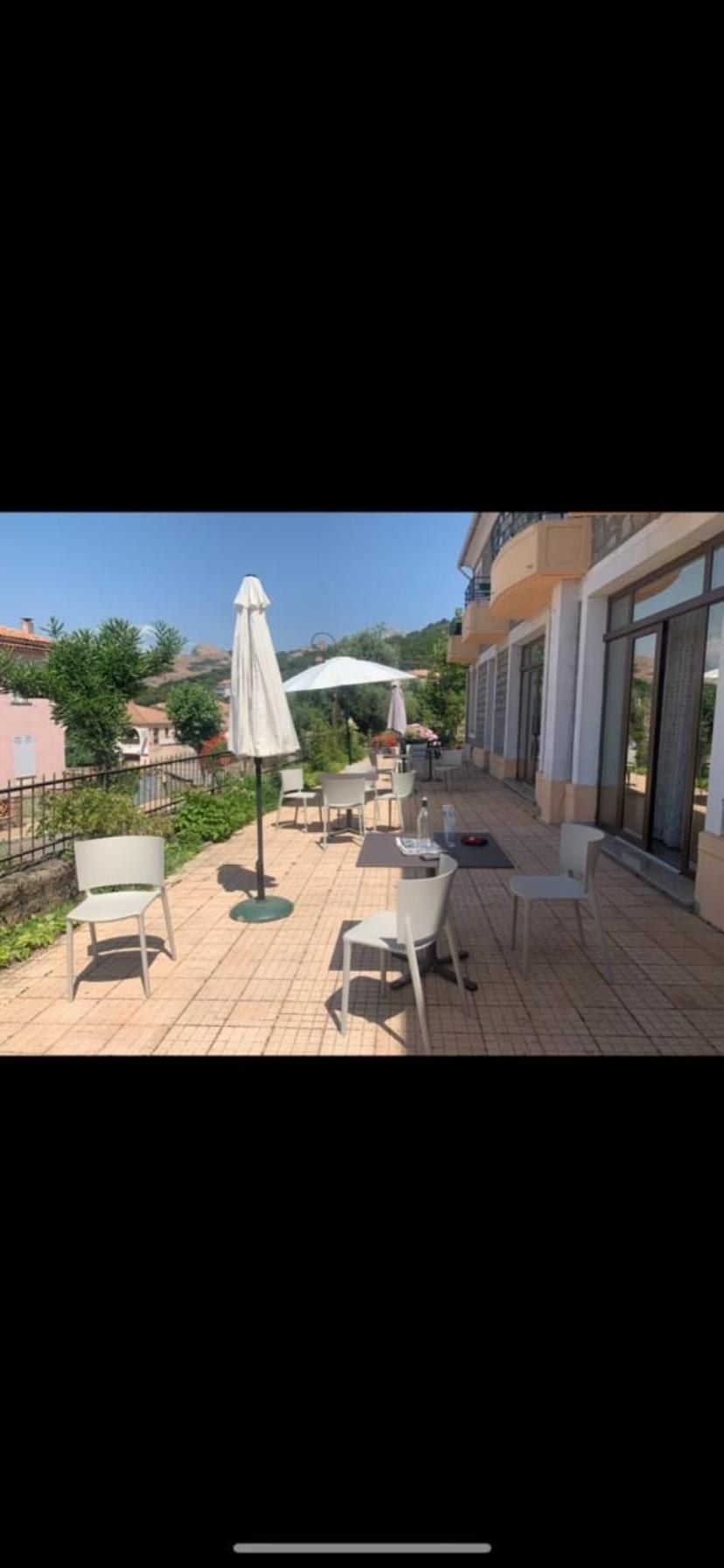 White patio furniture and folded umbrellas dot this sun-drenched terrace, where Mediterranean rooftops unfold below under brilliant blue skies and gentle coastal breezes.