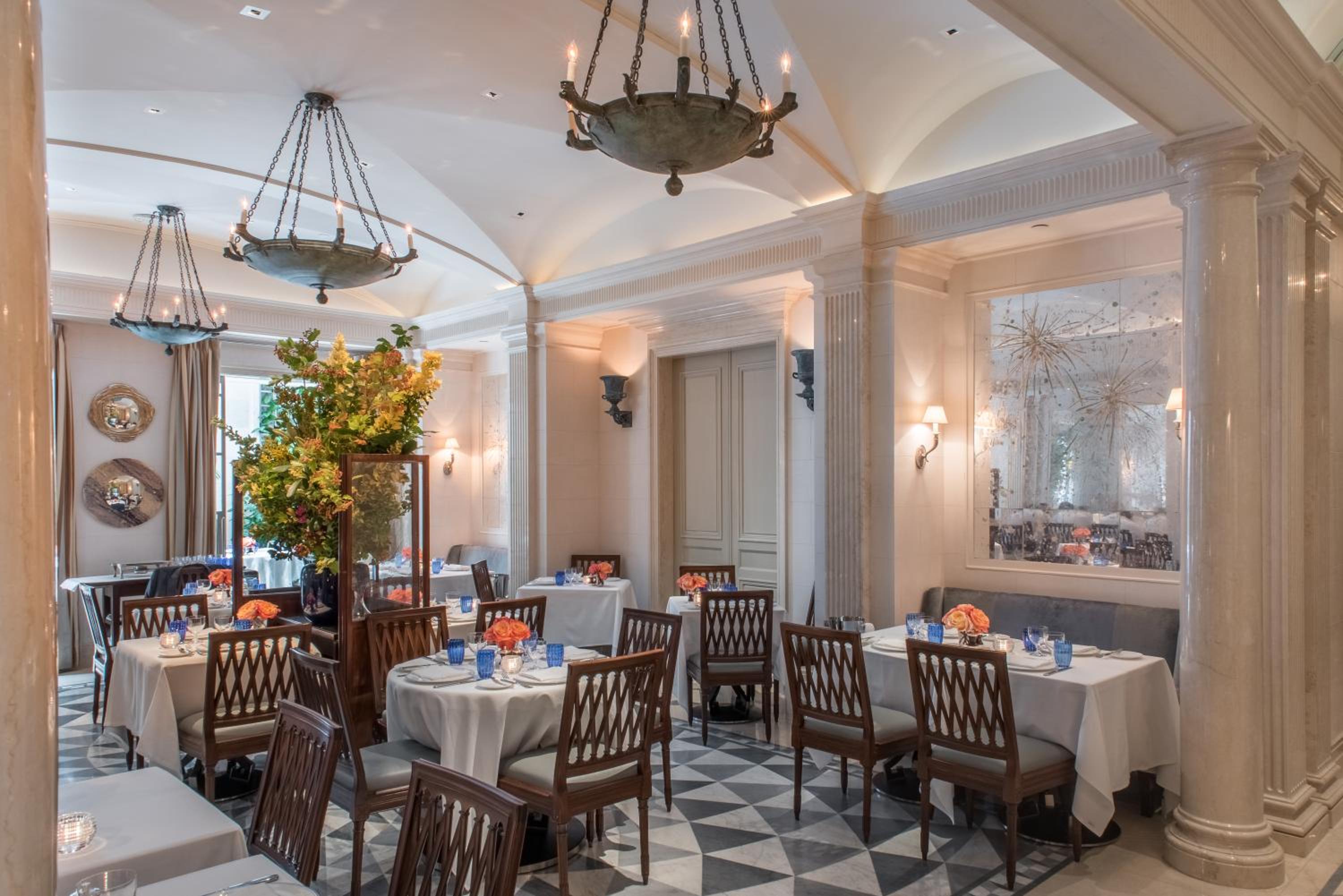 Crystal chandeliers cast a warm glow over crisp white linens and blue glassware in this intimate dining room, where coffered ceilings and classical columns create sophisticated elegance.