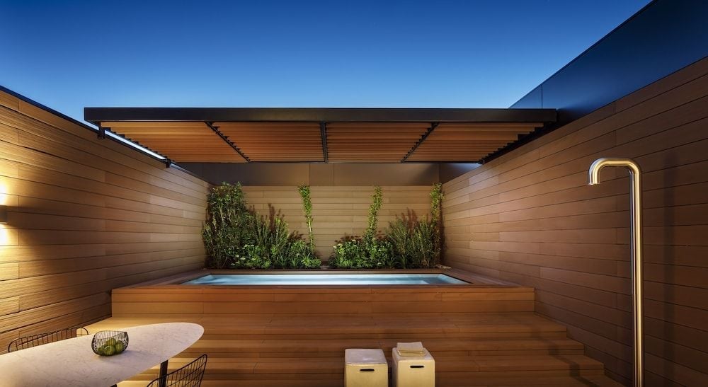 Warm timber decking frames an intimate rooftop spa pool, where sculptural planters and golden lighting create a serene urban sanctuary beneath the twilight sky.