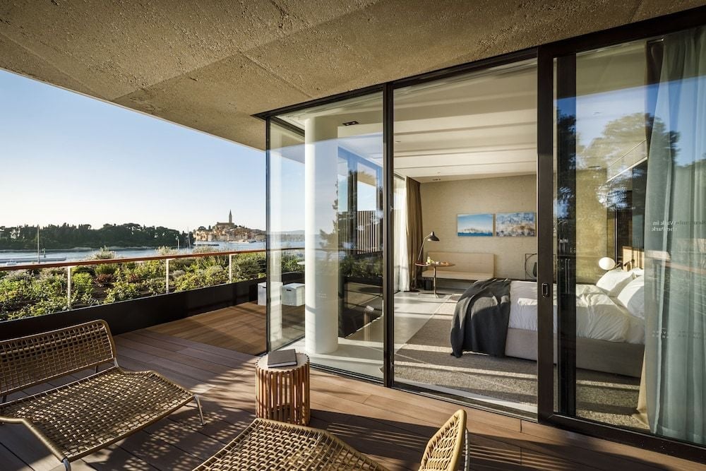 Floor-to-ceiling windows dissolve boundaries between bedroom and terrace, where wicker chairs frame panoramic vistas of Rovinj's medieval spires and Adriatic coastline.
