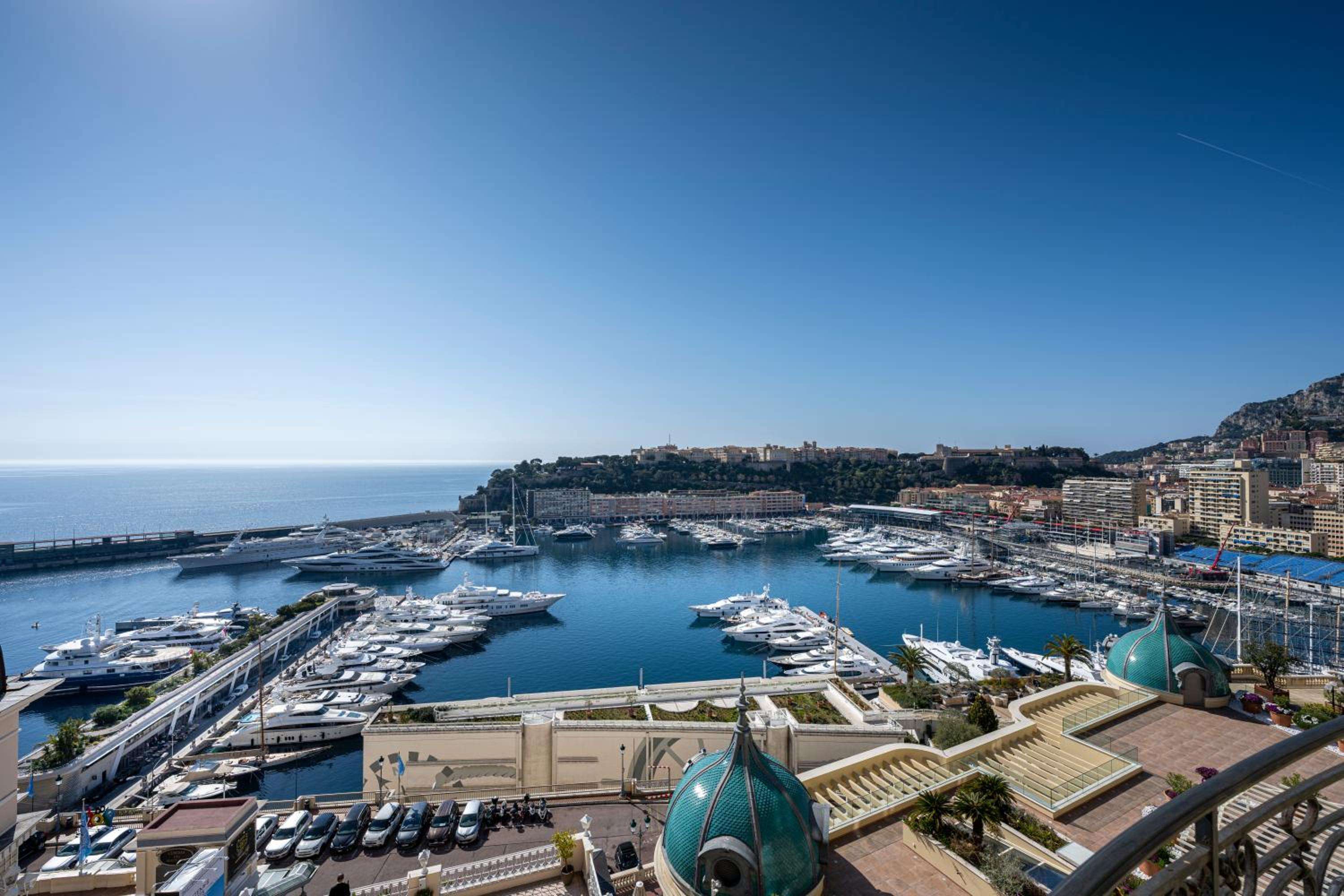 Monaco's legendary harbor unfolds below, where superyachts cluster like floating palaces against azure waters and the principality's terraced skyline climbs toward cloudless Mediterranean skies.