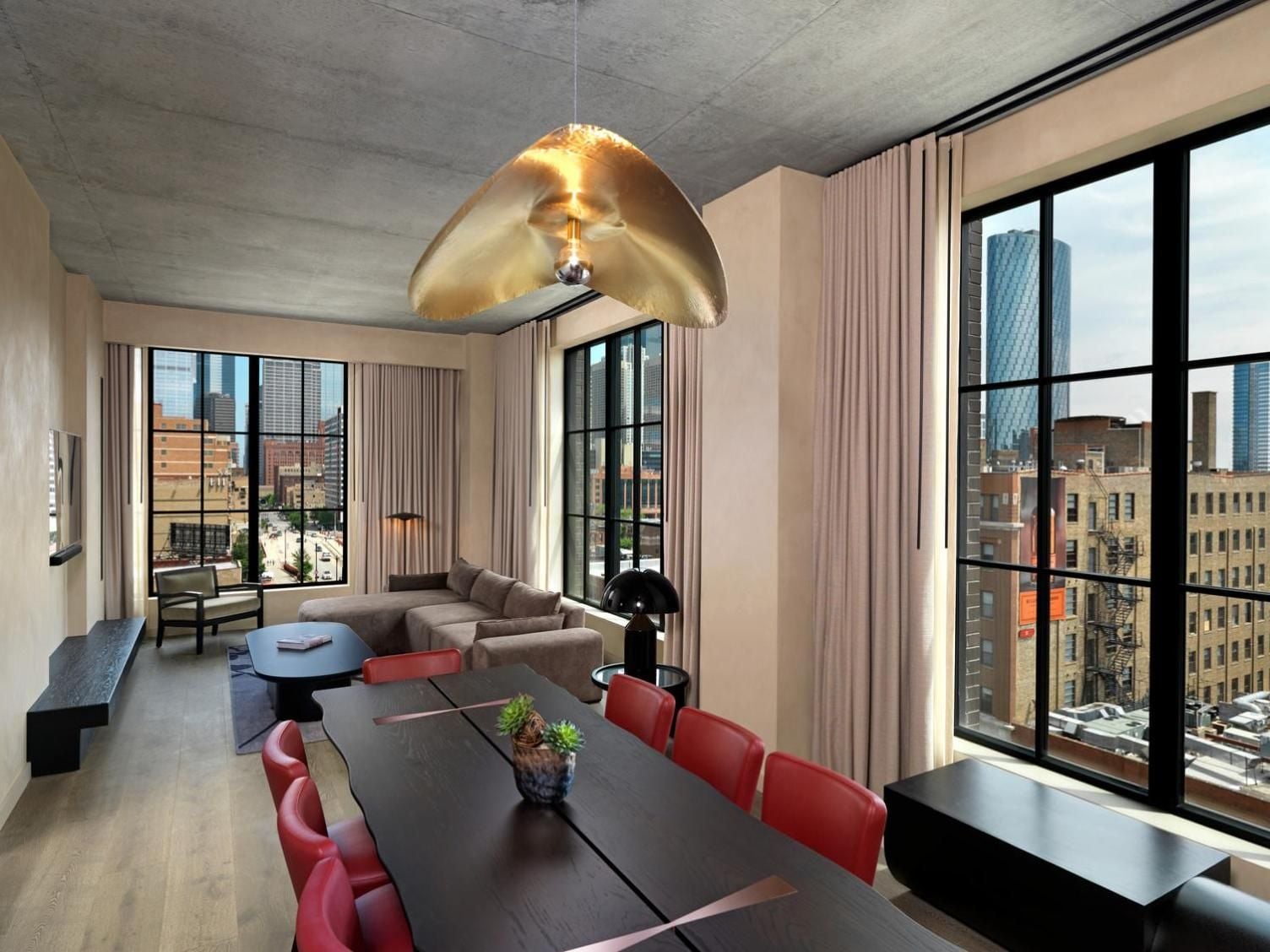 Floor-to-ceiling windows frame Chicago's skyline from this corner suite, where a sculptural gold pendant light casts warm shadows across the exposed concrete ceiling and coral-red dining chairs.