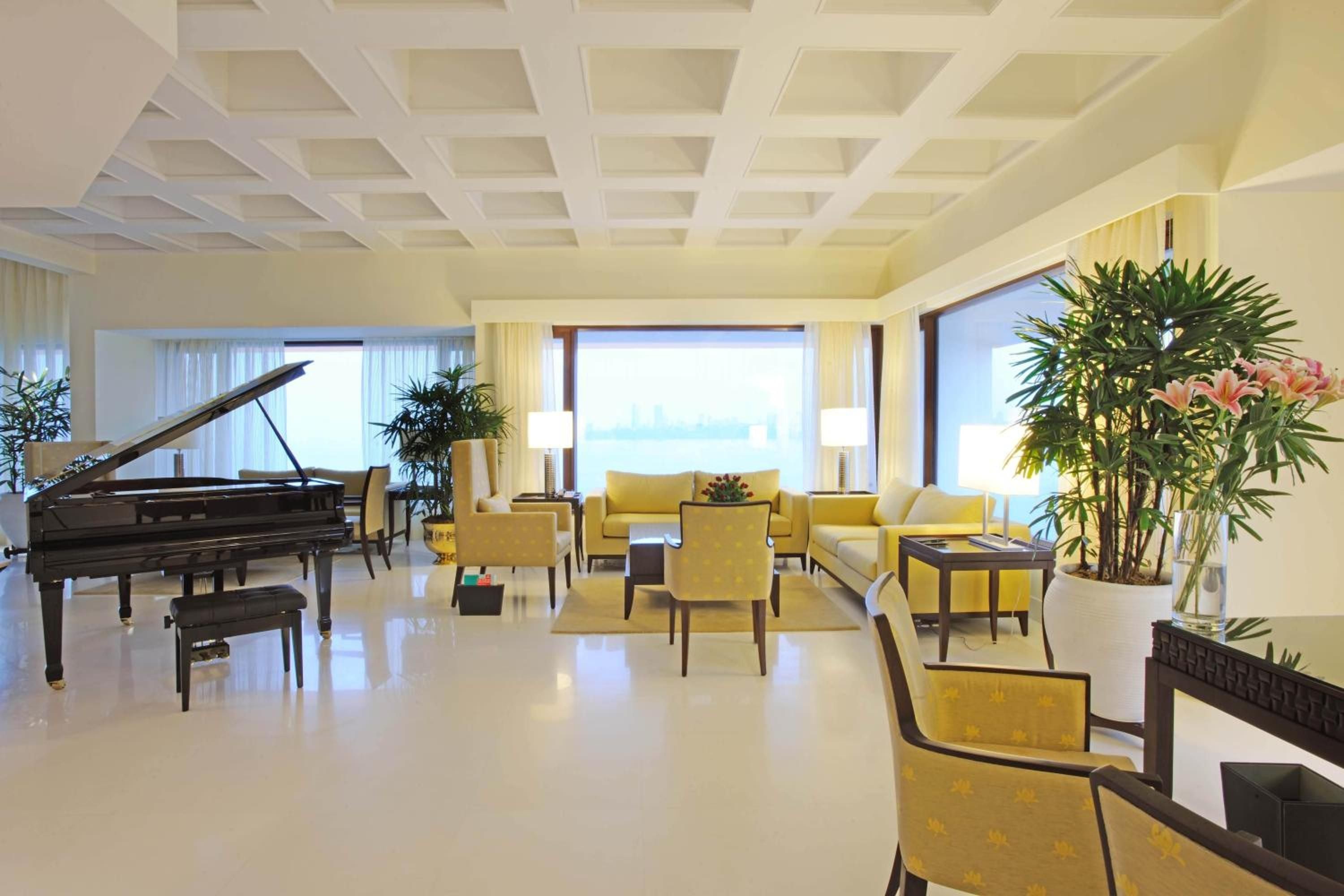 Golden afternoon light filters through floor-to-ceiling windows, illuminating the coffered ceiling and gleaming grand piano where yellow silk armchairs invite leisurely conversation.