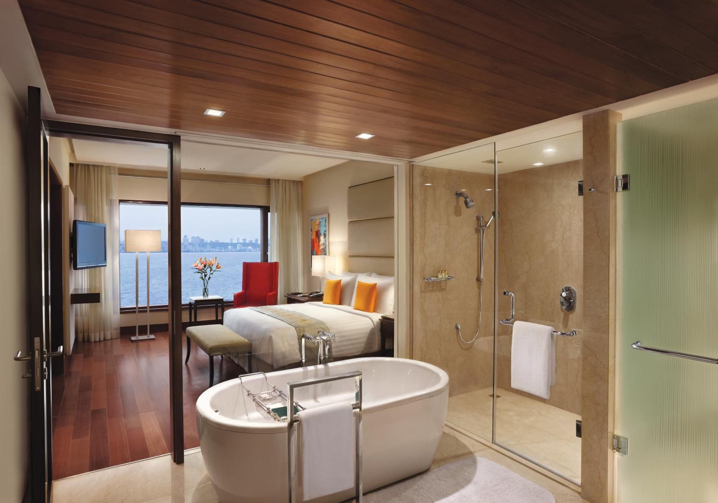 Floor-to-ceiling windows frame azure waters while a freestanding soaking tub anchors this sophisticated suite, where warm wood ceilings meet marble luxury.