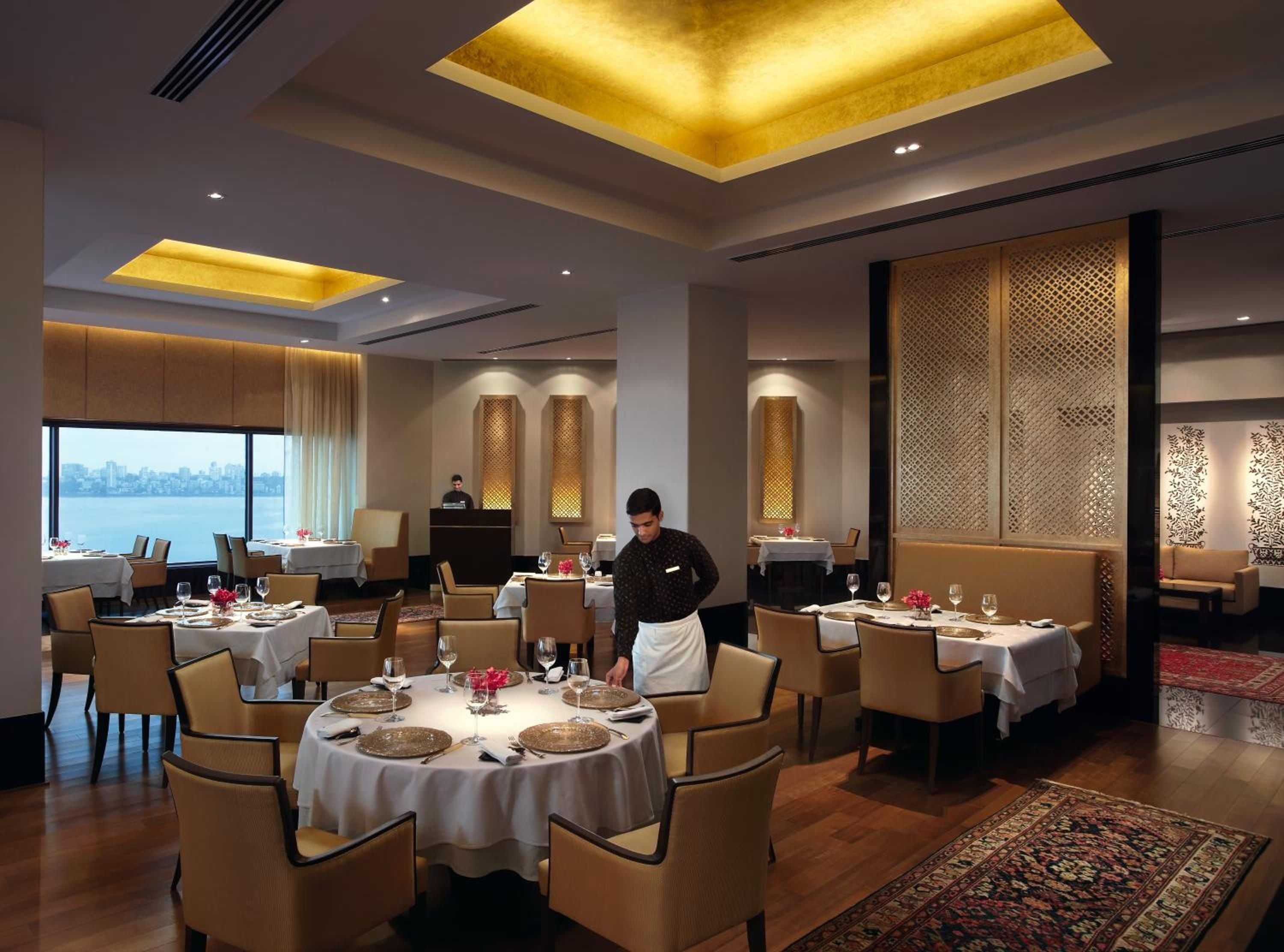 Golden ceiling coffers cast warm light over intimate tables while servers glide across polished floors, creating an atmosphere of refined elegance in this sophisticated dining room.