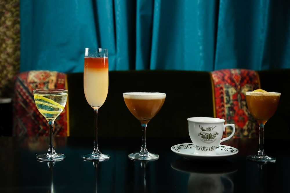 Five expertly crafted cocktails and coffee line the polished bar top, each telling its own story through layered hues and artful presentation against jewel-toned velvet seating.