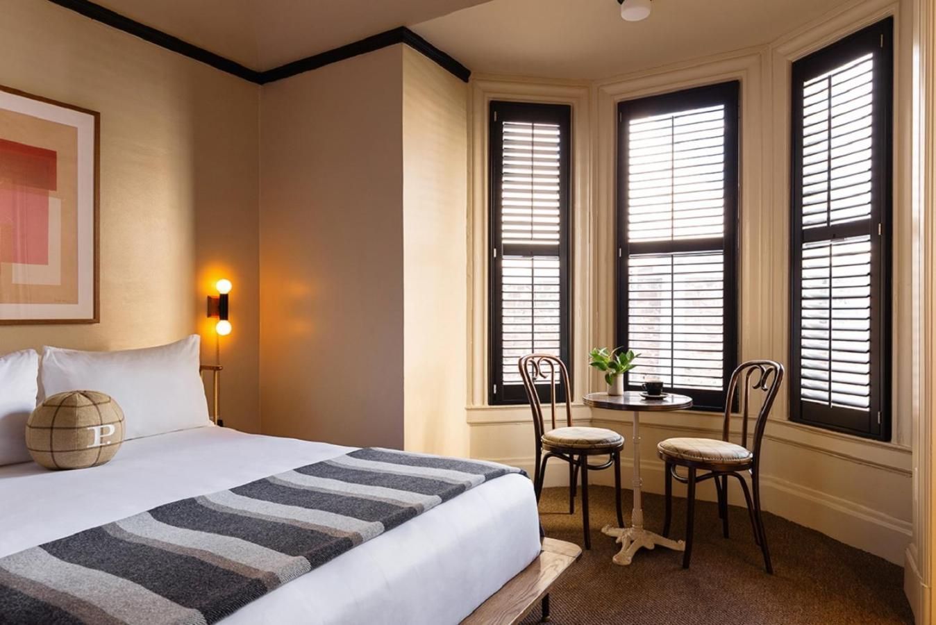 Warm amber light glows against taupe walls while bentwood chairs frame bay windows with dark shutters, creating an intimate corner perfect for morning coffee.