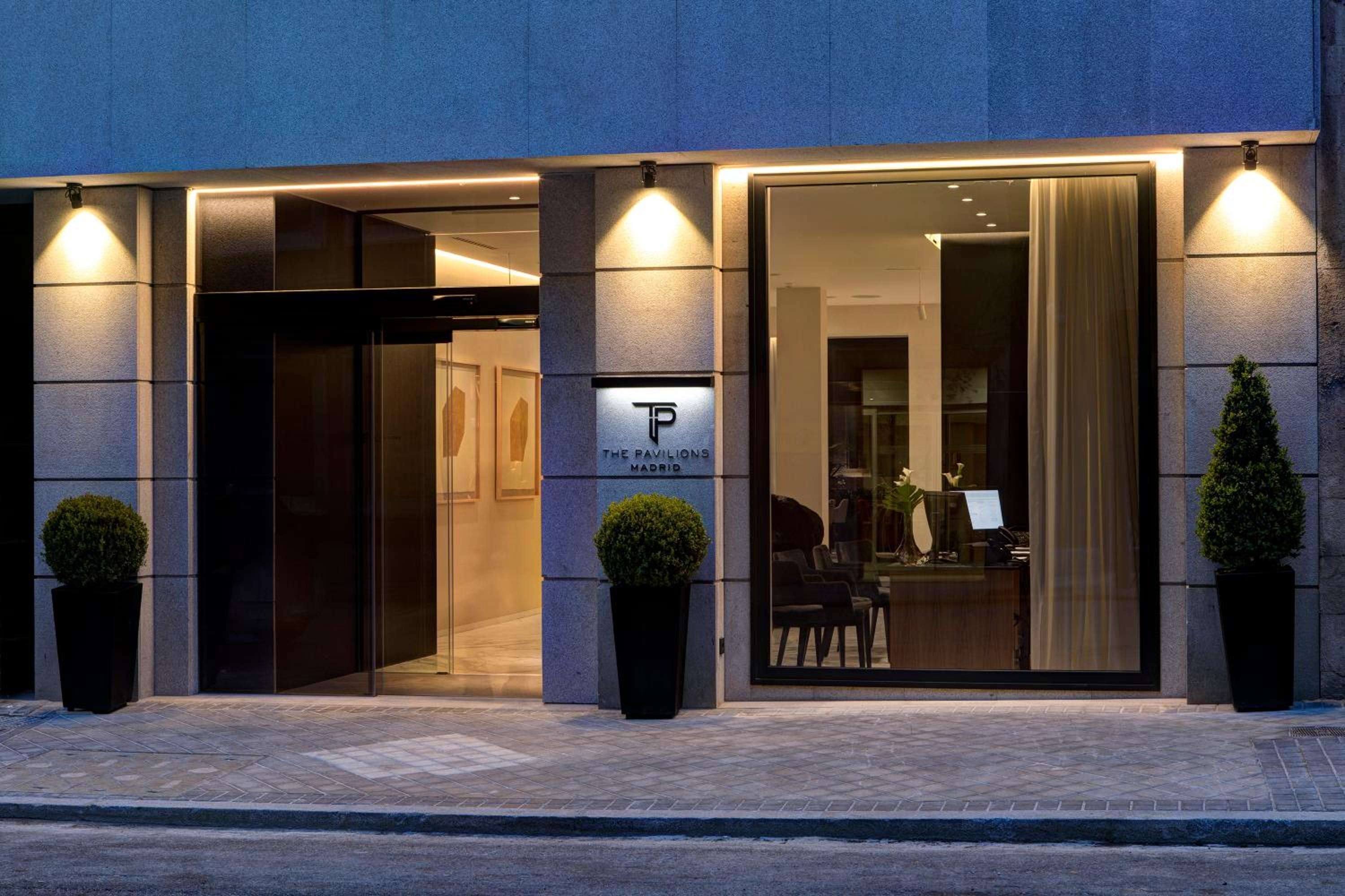 Warm golden light spills from The Pavilions Madrid's sophisticated entrance, where sleek stone columns and manicured topiaries create an inviting urban sanctuary.
