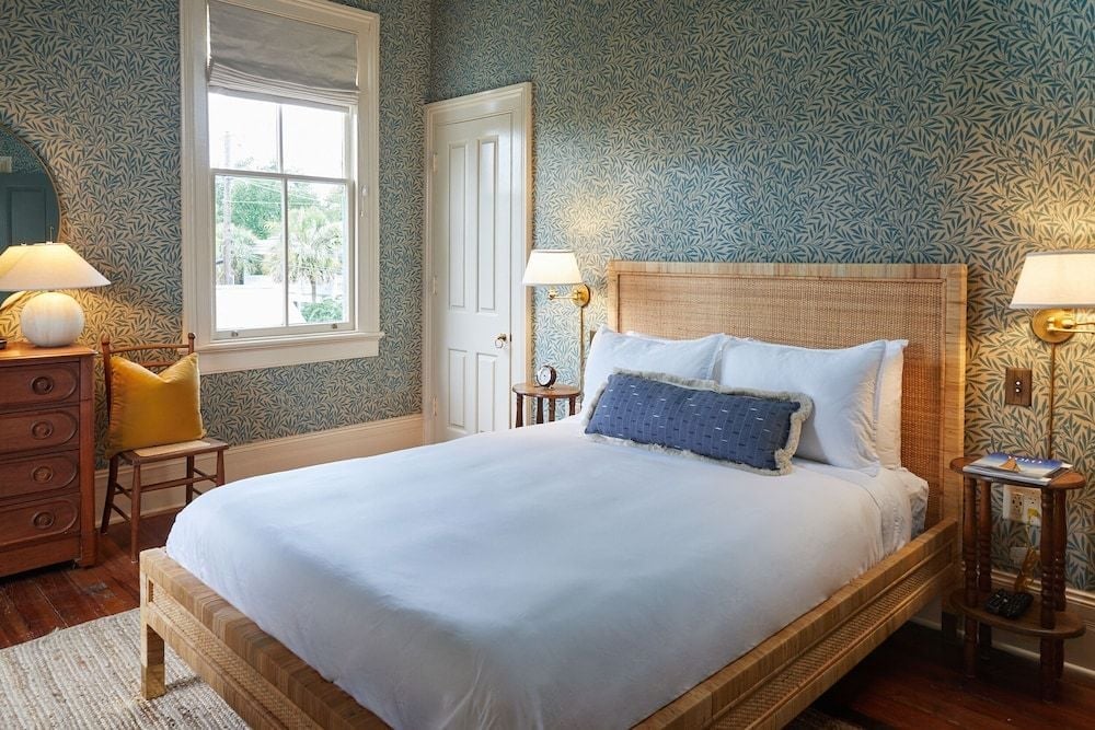 Botanical wallpaper and warm rattan create an intimate retreat, where morning light filters through vintage-style windows onto crisp white linens and polished hardwood floors.