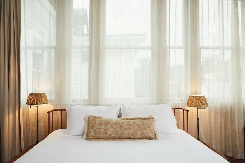Natural light floods through tall windows, illuminating crisp white linens and woven textures that create an intimate sanctuary above the bustling city streets.