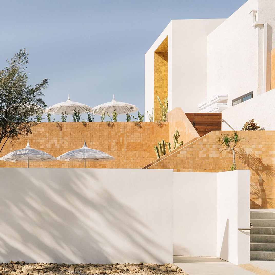 Geometric white forms and warm sandstone create striking contrasts, while striped umbrellas cast playful shadows across this modernist desert retreat's terraced poolside sanctuary.